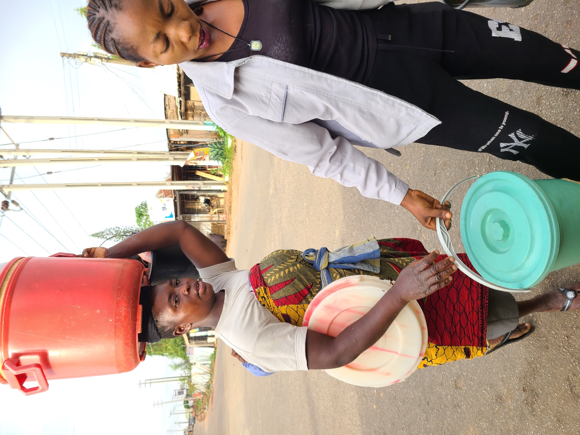 Two women are standing on a paved road, seemingly in the city of Jos, Nigeria. One woman is carrying a large red container on her head and a white container in her hand, while wearing a colorful wrap around her waist. The other woman is wearing black pants, a black top, and a light gray jacket, and she is holding a green bucket. The background shows buildings and utility poles along the road.