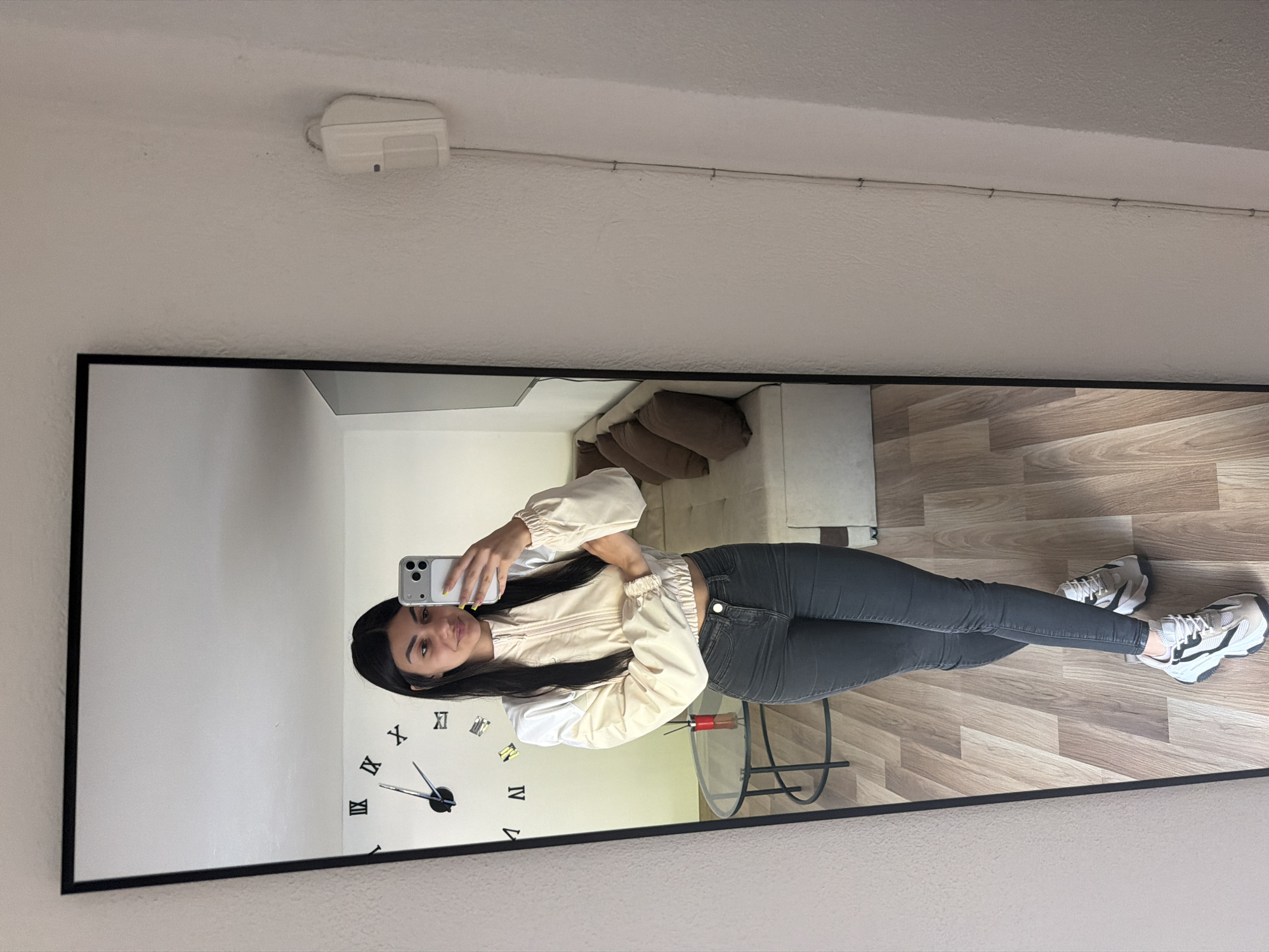 A mirror in a room reflects a young woman taking a selfie. She has long black hair and is wearing a light-colored jacket and dark jeans. The room reflected in the mirror contains a clock, a table, and flooring resembling wood planks. The top of the image shows a portion of the ceiling with a white device attached. The image provides no information to determine if this image was taken in Gabrovo, Bulgaria.
