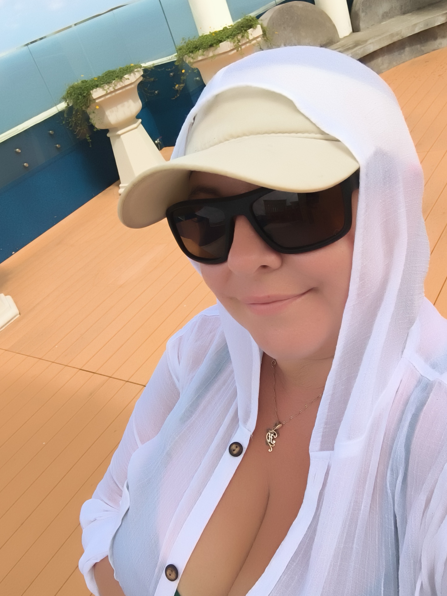 A woman, viewed from the chest up, is taking a selfie outdoors. She is wearing a white hooded cover-up, a tan baseball cap, and dark sunglasses. A gold necklace with a key charm is visible around her neck. The background shows a blue railing, a wooden deck, and some greenery. The scene appears to be on a cruise ship deck or a waterfront location. The exact location in Phú Quốc, Vietnam is not discernible from the image.
