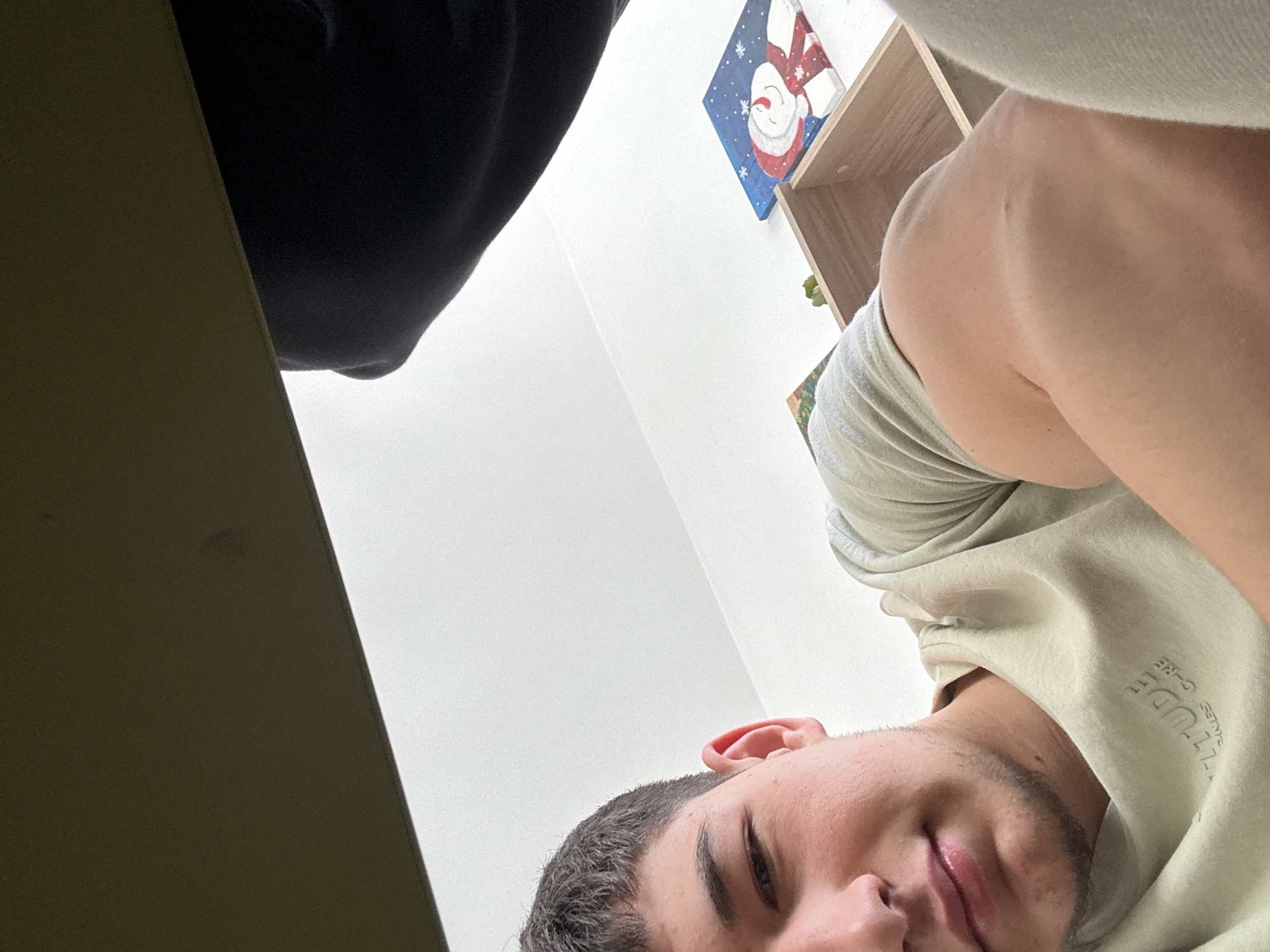 A young man is captured in a low-angle, indoor close-up shot, likely taken in Zlatograd, Bulgaria. His head is tilted to the left, and he looks towards the camera with a slight smirk or relaxed expression. He has short, dark hair and prominent eyebrows. He is wearing a light-colored (possibly beige or light green) t-shirt, with visible text on the right shoulder area that reads "ATTITUDE" and "SCALES G-CRE". A dark, possibly black, garment occupies the upper left corner of the frame.
The background consists primarily of a plain white wall. In the upper right corner, a light brown wooden shelf unit is visible. Above the shelf, a blue-framed picture or painting of a smiling snowman wearing a red hat and scarf, surrounded by white snowflakes, suggests a festive or winter theme. A dark olive green or brown surface is present in the lower left, possibly a piece of furniture or a table. The bright, even lighting indicates a well-lit indoor environment, likely during daytime. The unusual low perspective suggests the photo was taken from below the subject, who might be lying down or leaning back.