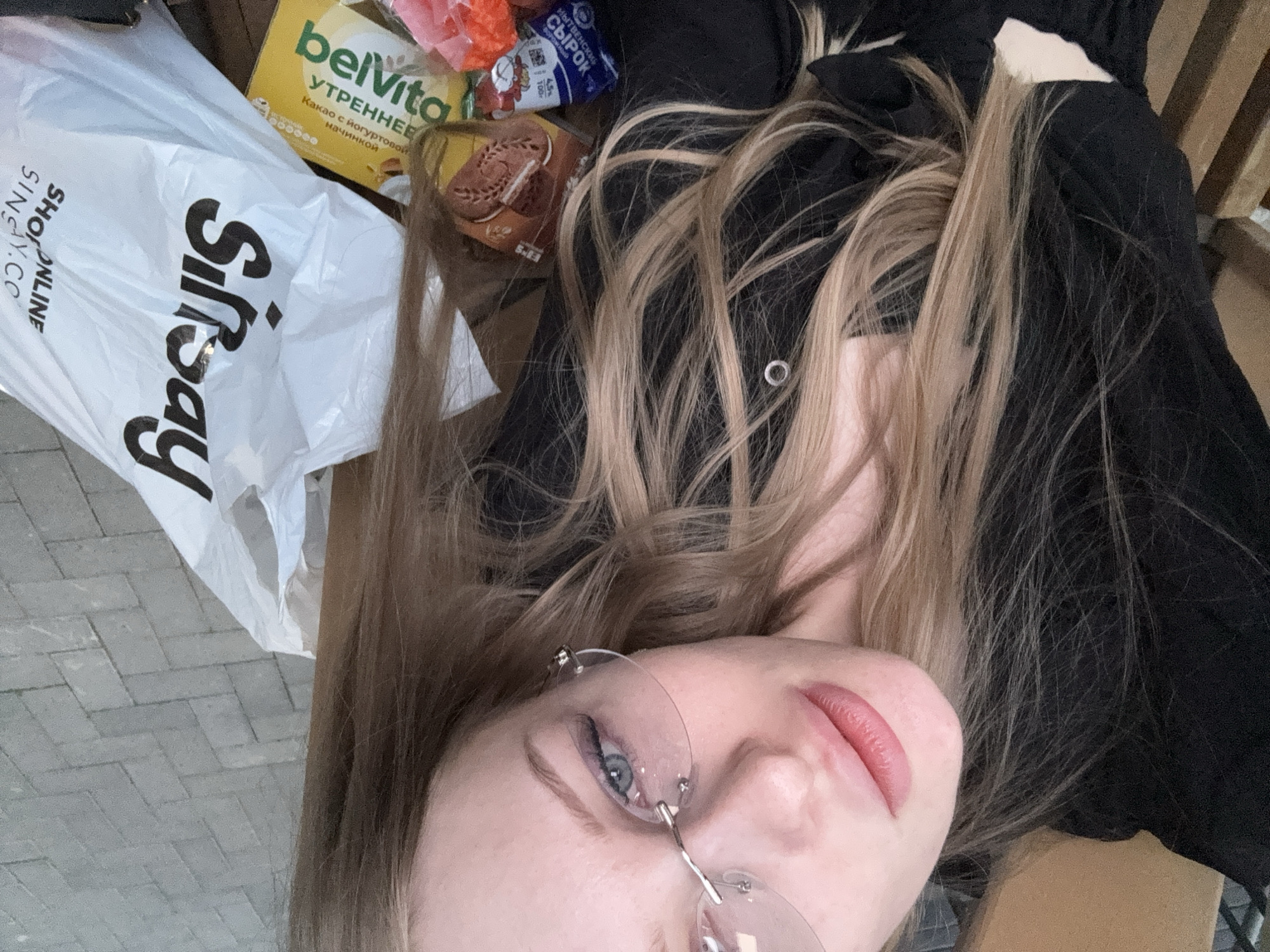A selfie shows a young woman with blonde hair and blue eyes wearing rimless glasses. Lying on what appears to be a wooden bench, she has a black top with a visible button. A white Sinsay shopping bag is next to her, along with food items including Belvita crackers and a dairy product labeled "сырок." The ground appears to be paved with gray bricks. The location is possibly in Perm, Russia, given the Russian text on the food products.