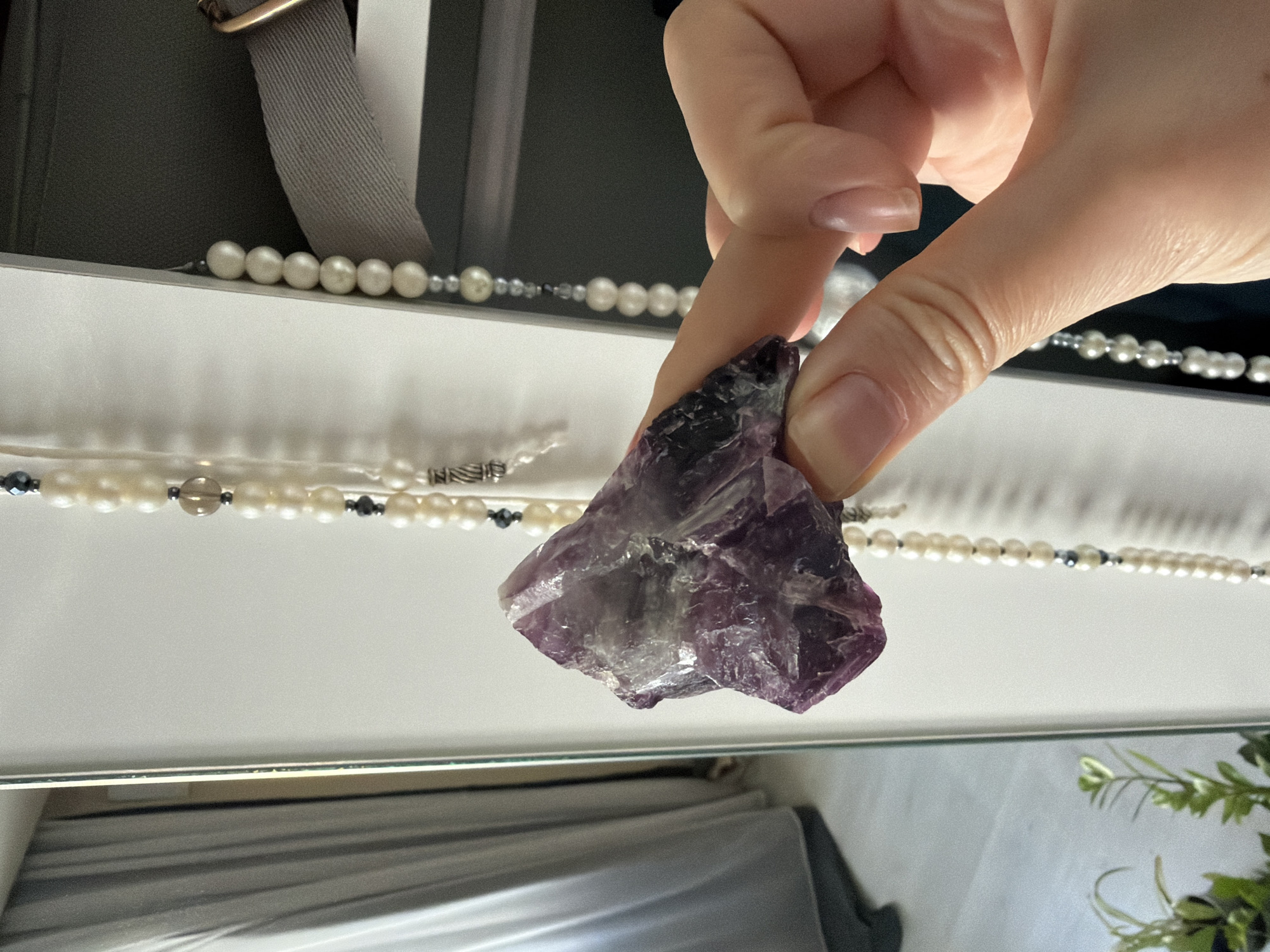A person's hand, with light skin tone and manicured nails, holds a purple, crystalline rock formation in front of a blurred background. The rock is multifaceted and appears to be amethyst, with lighter and darker purple hues. Behind the rock, several strands of pearl necklaces are visible, with occasional darker beads interspersed. The necklaces hang vertically, suggesting they are being held or displayed. To the left of the hand and rock, a large mirror reflects a curtain and a potted plant, indicating an indoor setting. The overall environment appears to be a well-lit room, possibly a closet or dressing area, given the presence of the necklaces and the reflective surface. There is no specific indication of the city of Izmir, Turkey.