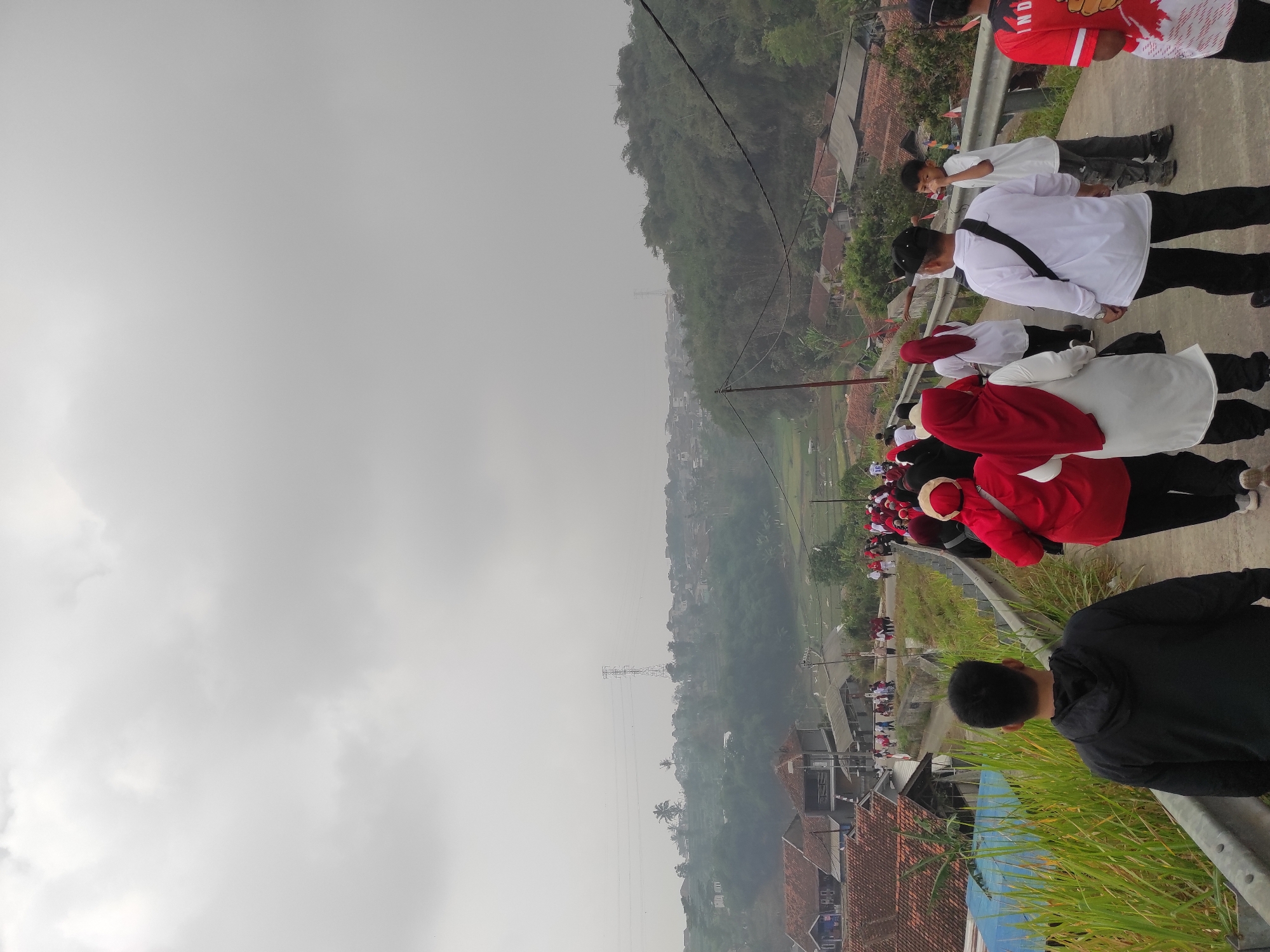 A group of people are walking up a paved road in what appears to be a rural or semi-rural area of Sumedang, Indonesia. The sky is overcast, and a hazy atmosphere obscures the distant landscape, which includes hills, scattered houses with red-tiled roofs, and some greenery. Some of the people are wearing red and white attire, possibly in observance of a national holiday. The road has a metal guardrail on the right side. In the foreground, a person in a black hoodie is walking away from the camera, and a few individuals in red and white are visible ahead on the road. The scene suggests an outdoor gathering or a community event.