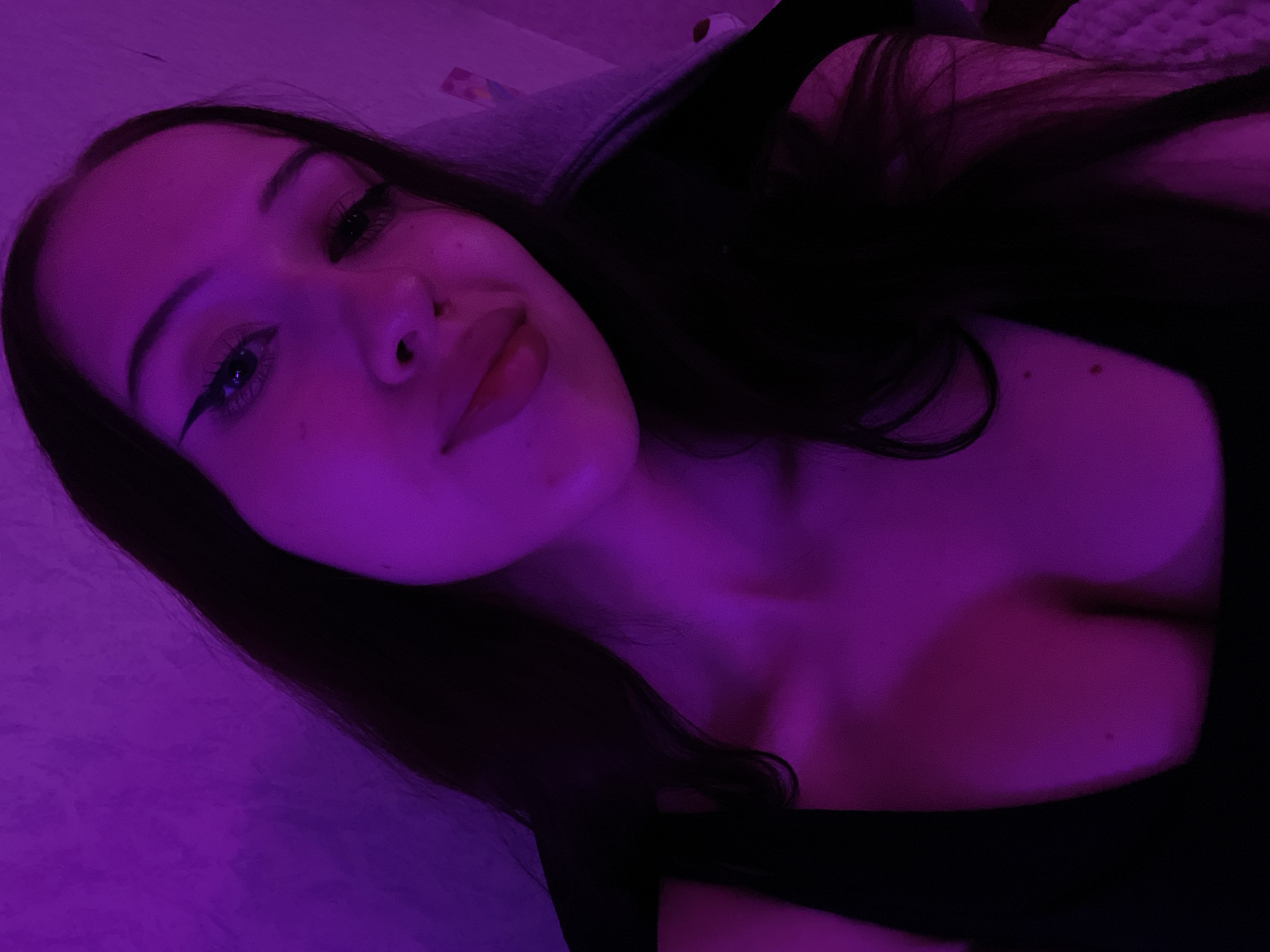 The image shows a close-up, waist-up selfie of a young woman indoors, under purple lighting. She has long dark hair, is wearing a black top, and has winged eyeliner. The background appears to be a wall with a textured surface, and a hint of a bed or couch with textured fabric is visible. The scene does not provide any information to suggest a location in Votkinsk, Russia.