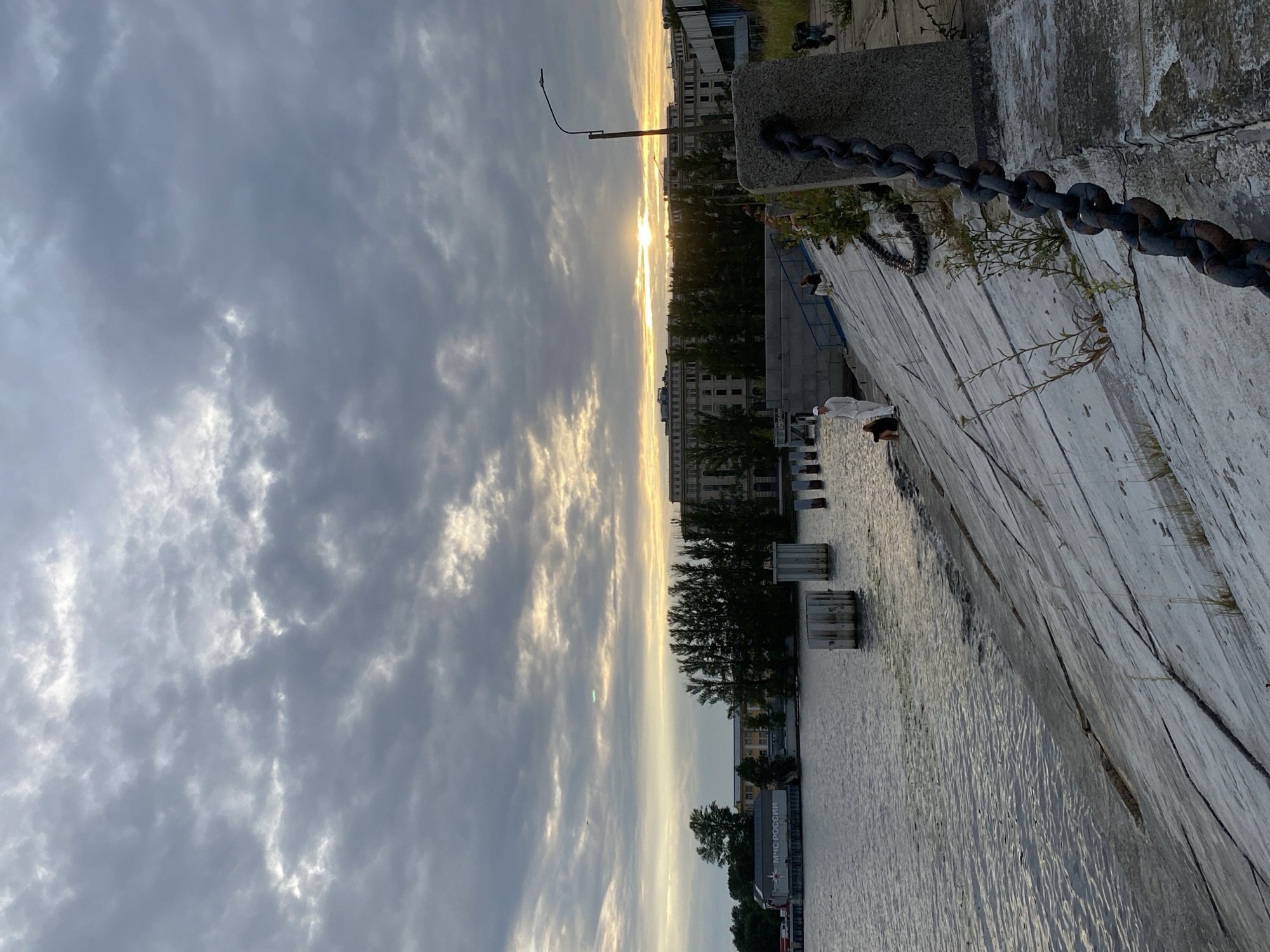 The image shows a wide shot of a riverfront area in Saint Petersburg, Russia, at sunset. The sky is filled with dramatic, dark clouds interspersed with patches of bright sunlight breaking through, indicating the sun is low on the horizon. The river, which appears to be the Neva River, occupies the left half of the frame, its surface rippled by a gentle breeze.
Along the riverbank, a concrete embankment slopes down to the water. Several people are visible on the embankment. In the foreground on the right, a thick, rusty chain is attached to a concrete post, stretching down the embankment. A lamppost is also visible on the right.
Across the river, a long, multi-story building with many windows is situated behind a line of tall trees. On the far left, a building with the inscription "МЧС РОССИИ" (Ministry of Emergency Situations of Russia) is visible. In the water, there are several concrete pillars or structures. The overall atmosphere is serene, with the fading light creating a somewhat moody and dramatic ambiance.
