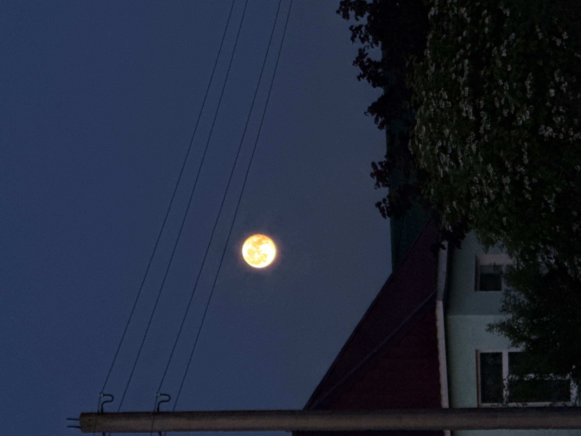 The image displays a night or late dusk scene, featuring a prominent, bright, yellowish-orange full moon against a dark blue sky. Several thin, dark utility lines stretch diagonally across the sky from the lower left, passing in front of the moon and extending towards the upper right. These lines are attached to a dark, horizontal utility pole or crossarm visible at the bottom left of the frame, which features distinct metal fixtures on its top edge.
On the right side of the image, parts of buildings and dense foliage are visible. A building with a dark red or maroon sloped roof occupies the mid-right. Adjacent to or beneath this roofline, a lighter-colored, possibly light green or blue, section of a building is seen, featuring at least two windows: a smaller square one above and a larger rectangular one below. Dark, leafy trees or bushes frame the far right and top right of the image, with some small white elements that could be blossoms or highlights on leaves. The scene depicts an outdoor setting, likely in a semi-urban or rural area of Лабжанскі сельскі Савет, Belarus. No people or text are visible.