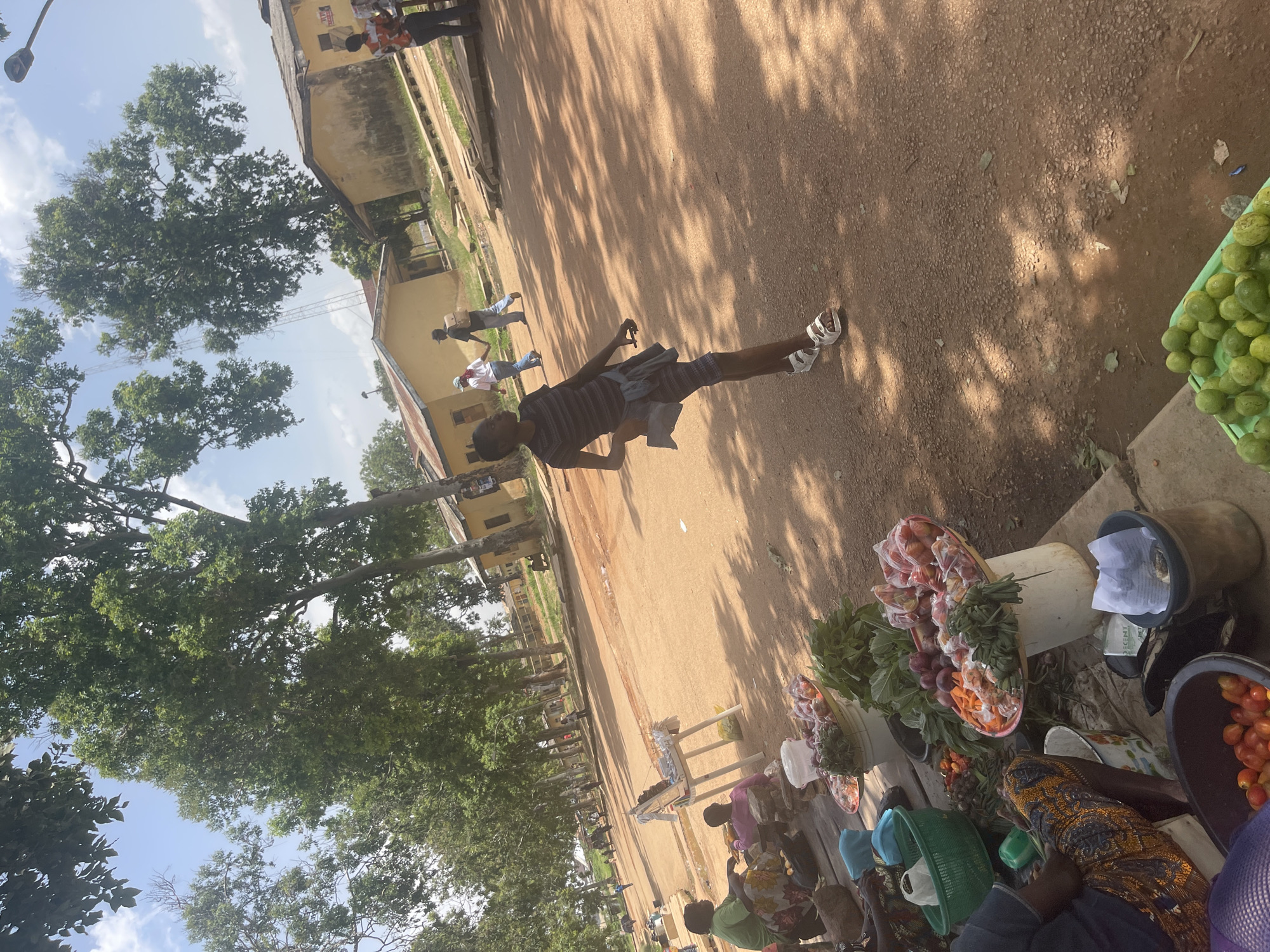 In Rafin Par, Nigeria, a group of people are gathered at what appears to be a market. The scene is set outdoors on a dirt path, with buildings and large trees lining one side. The time of day is likely mid-morning or afternoon, indicated by the dappled sunlight and shadows cast by the trees. The weather seems pleasant, with a clear blue sky visible through the leaves.
The central subject is a young man wearing a striped t-shirt and checkered shorts, walking with a slight swagger. He is holding a grey garment over his arm and wears white sandals. Behind him, several other individuals are visible in the distance, some walking and others standing.
In the foreground and to the right of the image, a market stall is set up. Various fruits and vegetables are displayed in baskets and containers. We see what look like tomatoes, onions, and leafy greens. Some people are seated behind the stall, appearing to be vendors or shoppers. One person is wearing a colorful patterned dress.
The overall atmosphere suggests a lively, everyday market scene. The presence of both shoppers and vendors, the abundance of fresh produce, and the casual gathering of people point to a typical day of commerce and social interaction in this Nigerian community. The image is captured from a slightly tilted angle, adding a dynamic perspective to the scene. No legible text is visible in the image.