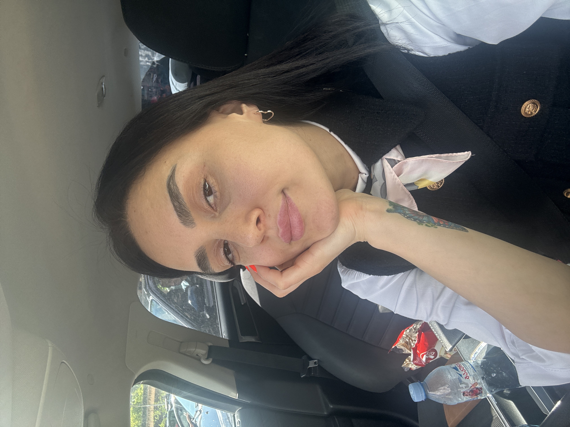 The image shows a young woman sitting in a car, likely as a passenger, posing for a close-up shot under bright daylight. She is looking towards the viewer with a gentle smile, resting her chin on her left hand. She has long, dark hair, prominent eyebrows, and wears light pink lipstick and red nail polish on her fingers. A small, heart-shaped earring is visible in her left ear.
She is dressed in a white collared shirt, layered with a black vest or jacket featuring decorative gold buttons. A light pink patterned scarf or tie is visible around her neck. A colorful tattoo, appearing to be a butterfly or floral design, is visible on her left forearm.
The car's interior is visible around her, including dark seats, a seatbelt, and parts of the beige ceiling and window frames. On the car's console or floor, a plastic bottle of "BON AQUA" water with a blue cap and a crumpled red and gold snack bag are present. Through the car windows, blurred trees and reflections of other vehicles suggest an outdoor, urban or suburban setting. The context for the location is Tbilisi, Georgia.