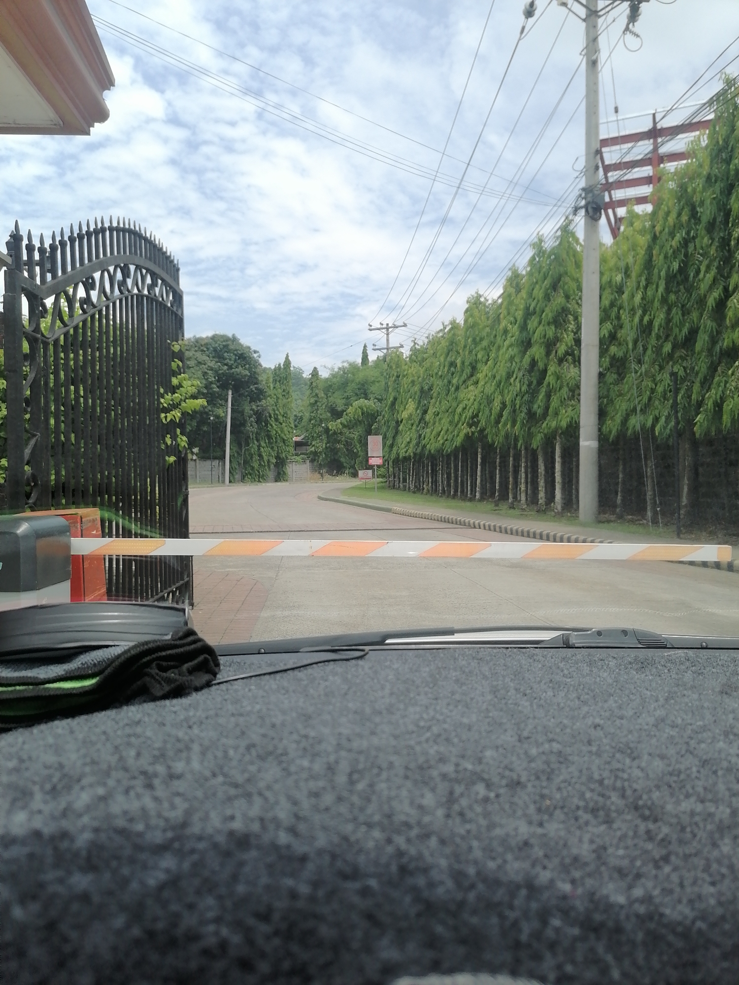 The image captures a view from inside a vehicle, looking out through the windshield at a gated entrance. The lower part of the frame shows the vehicle's dark, textured dashboard with a black and green cleaning cloth resting on it.
In the midground, a black, ornate metal gate is partially open on the left, revealing a paved road beyond. A white and orange striped barrier arm is lowered across the road, blocking entry. To the left of the barrier is its control mechanism. The road surface changes from brick paving near the gate to asphalt further in. A concrete curb with black and white stripes runs along the edge of the road, leading into the distance.
The background is dominated by lush greenery. On the right, a long line of tall, slender evergreen trees forms a dense natural wall alongside the road, extending far into the background. Utility poles with numerous overhead power lines are visible against the cloudy sky on the right side. Behind the trees and utility pole on the far right, a large red-beamed structure, possibly a building under construction, can be seen. On the left side of the road, further down, another collection of trees and foliage is visible. A red circular sign with "30" in white is faintly visible on the left side of the road in the distance, indicating a speed limit. The sky is partly cloudy with white clouds.
The scene depicts a controlled access point, likely to a private property, resort, or residential community in Davao City, Philippines. The time of day appears to be daytime, under an overcast or partly cloudy sky. The vehicle is stationary, waiting at the closed barrier.