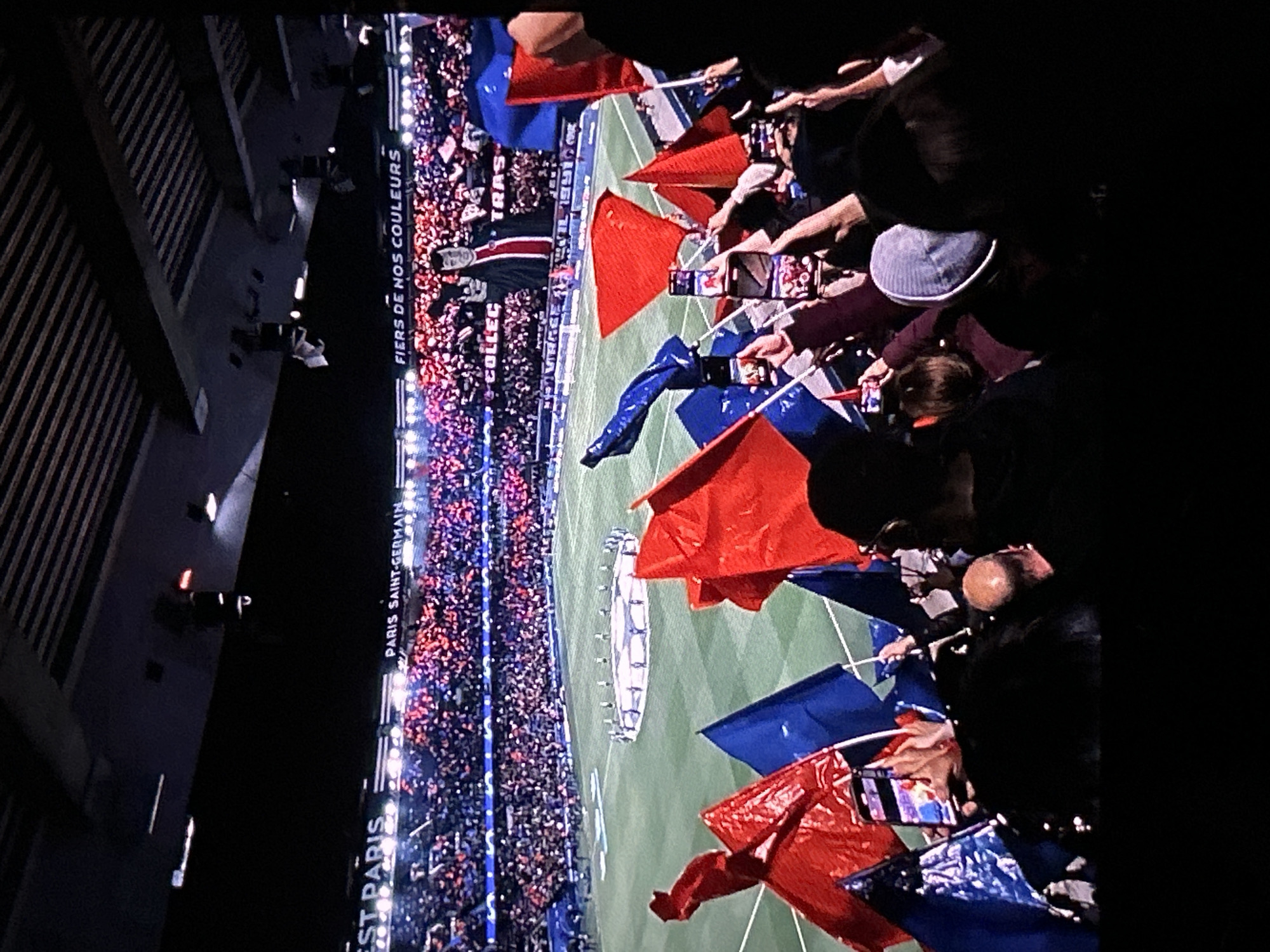 The image displays a vibrant nighttime scene inside a large football stadium, viewed from an elevated perspective, possibly from an upper tier or balcony. A considerable crowd of spectators fills the stands, illuminated by bright stadium lights.
In the foreground, numerous individuals are visible, many holding up small red and blue flags. Several people are actively engaged in recording the event with their smartphones, holding them high to capture the action. The field below is a lush green, featuring a distinct large circular emblem or structure at its center, typical of pre-game or post-game ceremonies.
The overall setting indicates a sporting event or a major public gathering. The time of day is clearly night, evidenced by the darkness surrounding the artificially lit stadium. The atmosphere appears festive and energetic, with the crowd’s visible engagement and the prominent display of flags.
Crucial textual cues are present on banners within the stadium. Visible text includes "FIERS DE NOS COULEURS" (Proud of our colors), "COLLEC" (partially visible), and distinctly "PARIS SAINT-GERMAIN" along with "PARIS". These inscriptions strongly link the event to the Paris Saint-Germain Football Club, suggesting the location is in Paris, France, likely the Parc des Princes stadium. This information directly contradicts the provided location context of "Bania, Bulgaria."
