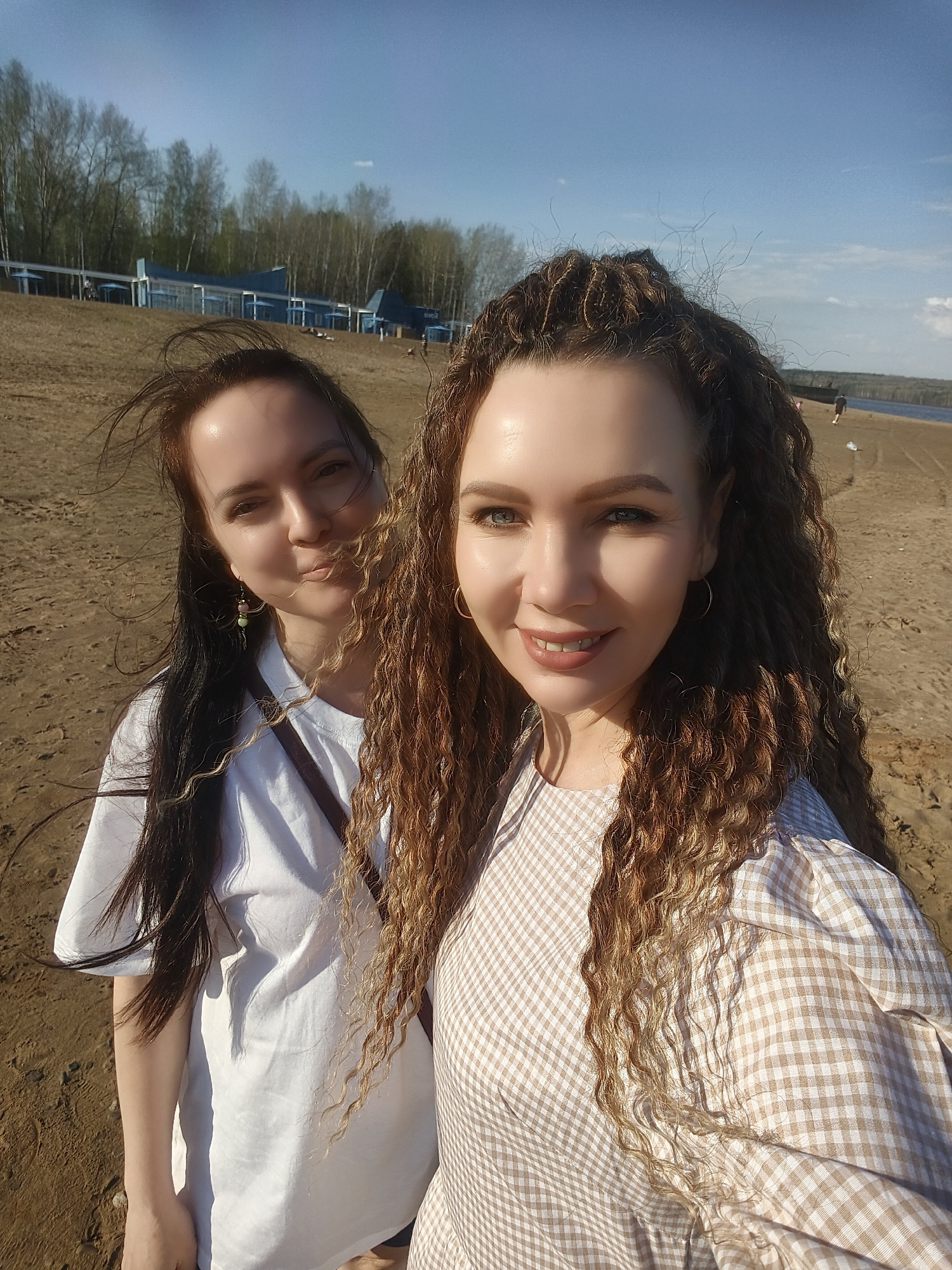 Two women are posing for a selfie on a sandy beach. The woman on the right has long, curly brown hair and is wearing a tan and white checkered dress. She has gold hoop earrings and is smiling at the camera. The woman on the left has long, straight dark hair and is wearing a white t-shirt. She is slightly behind the woman on the right and is making a playful face. The beach extends behind them, and in the distance, there are trees, some blue structures, and a body of water. The sky is blue with some clouds.