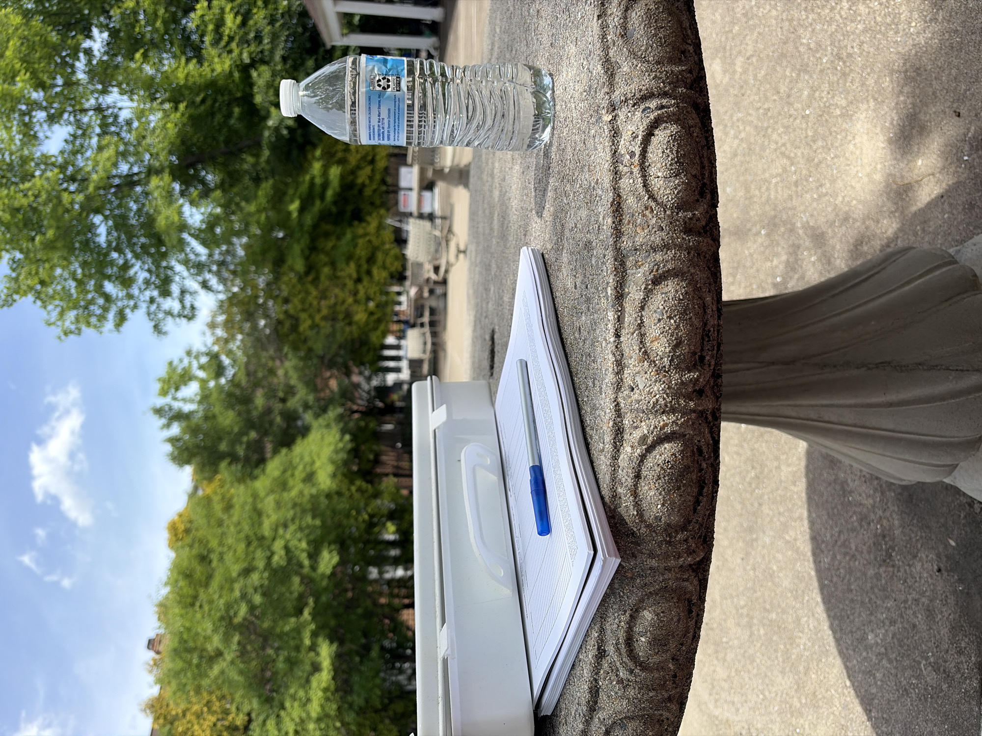 A water bottle sits on a round concrete table next to a white clipboard with papers and a blue pen on top. The table is located outdoors, with green trees and a light blue sky visible in the background. The OCR text on the water bottle includes "DISTRIBUTED BY: Wal-Mart Stores, Inc., AR 72716 PURIFIED BY: Reverse Osmosis PLASTIC BOTTLE". The location cannot be determined as being in Edison, New Jersey as there are no other clues for the location to be identified.