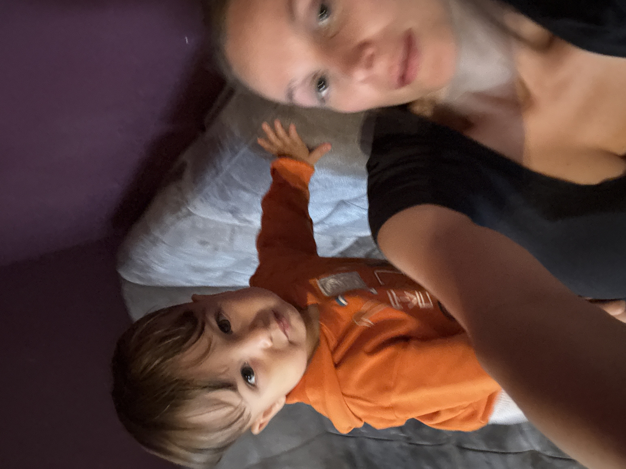 In this close-up, top-down selfie taken from the perspective of someone lying down, a young child with brown hair and brown eyes is the primary subject. The child is wearing an orange hooded sweatshirt with a graphic design on the front and appears to be reaching out towards the camera. To the right of the child, the face of an adult, likely female, is partially visible, with light-colored eyes and blonde hair. The adult's arm extends into the frame in the foreground, obscuring a portion of the image. The background consists of a textured gray surface, possibly a couch or bed, and a dark purple surface, likely a wall. The scene is dimly lit, suggesting an indoor setting. The photograph was taken in the city of Überherrn, Germany.