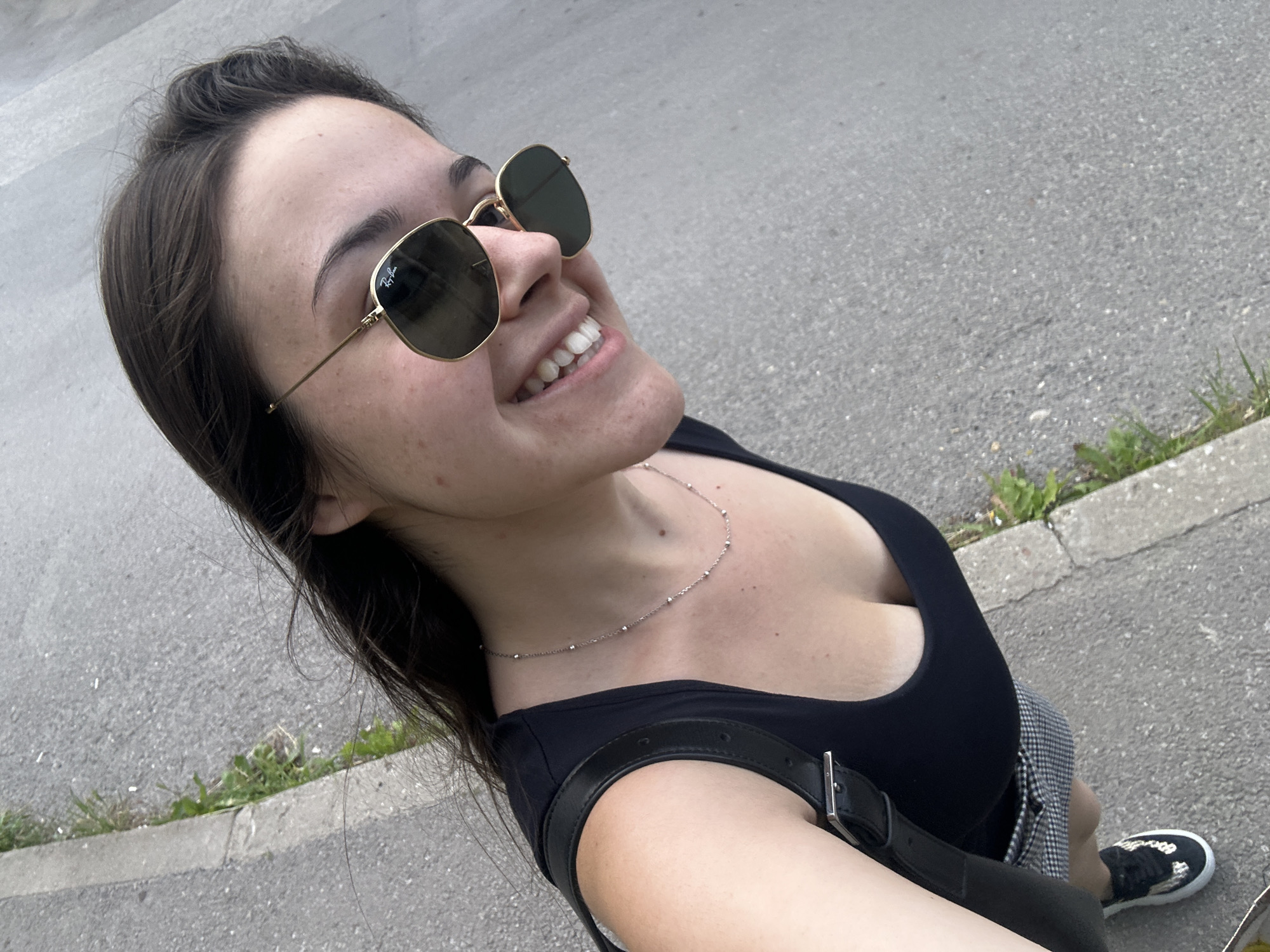 A young woman with long, dark hair and sunglasses is taking a selfie outdoors on a paved surface, possibly a sidewalk or street, with some sparse grass along the edge. She is wearing a black tank top, a checkered skirt, and black and white sneakers. She is smiling and looking towards the camera. The environment appears to be urban, with the textured gray asphalt taking up most of the background. The lighting suggests it is daytime. There are no specific landmarks visible that would definitively place the location within Sofia, Bulgaria.