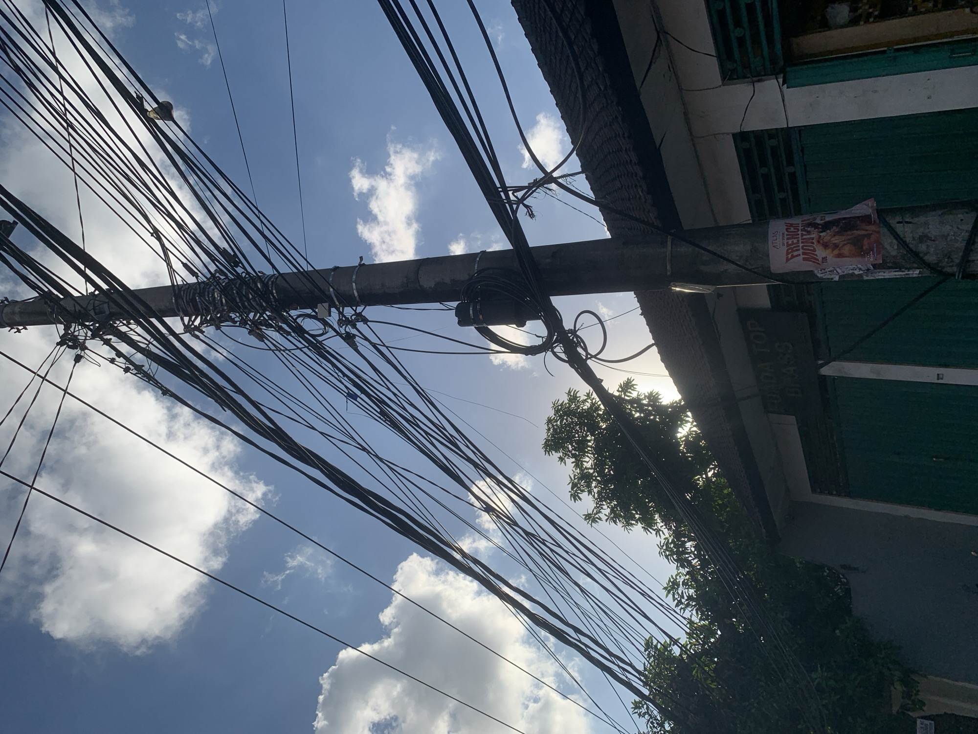 The image captures a street scene in North Kuta, Indonesia, dominated by a utility pole heavily laden with numerous electrical wires against a bright blue sky with scattered white clouds. The pole itself is a concrete structure, and the wires are tangled and crisscrossed, obscuring much of the sky. A small, dark electrical box is attached to the pole, with more wires bundled around it.
Below the wires and to the right of the pole, a dark-tiled roof of a building is visible. A portion of the building's facade shows green doors or shutters. A poster, partially torn and weathered, is affixed to the utility pole. It appears to be an advertisement, with the text "FRENCH MONTANA" and "ATLAS" clearly visible.
To the left of the pole, lush green foliage from trees is present, with some of the leaves silhouetted against the bright sky. The sunlight is strong, casting some glare and making it difficult to discern finer details in certain areas. The overall impression is one of a typical urban or semi-urban environment where overhead electrical infrastructure is prominent. There are no people visible in the image.