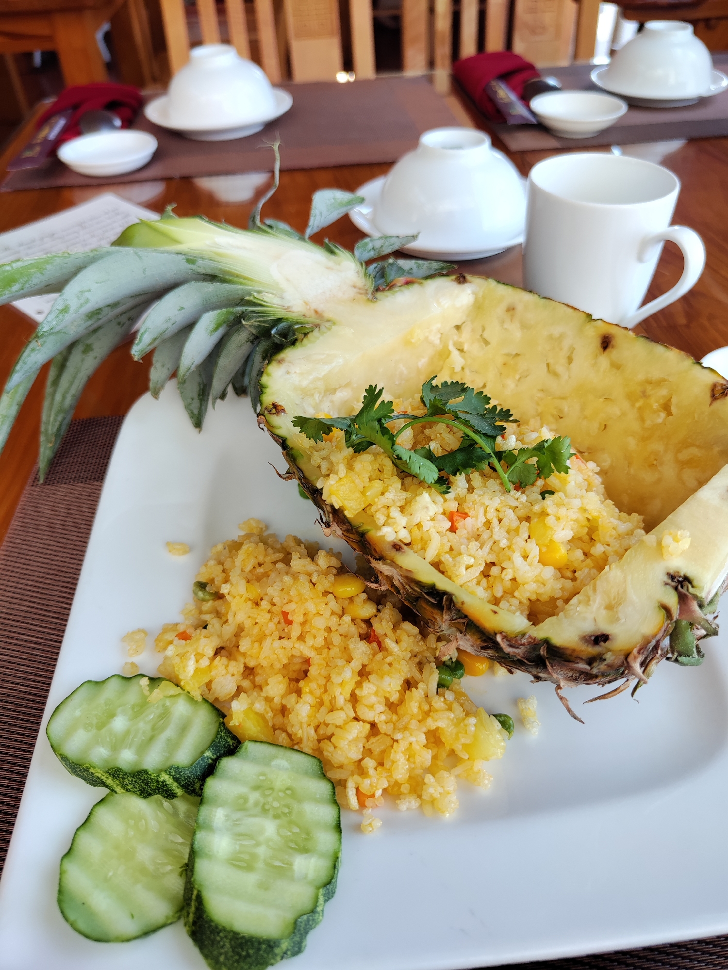 A close-up, high-angle view captures a dish of pineapple fried rice served on a light brown wooden table in Da Lat, Vietnam. The fried rice is presented inside a hollowed-out half pineapple, positioned diagonally on a large white rectangular plate. The pineapple retains its green leafy crown at the top left. The rice is yellowish, containing visible pieces of corn, diced carrots, and peas, and is garnished with fresh green cilantro sprigs. A small portion of the fried rice is scattered directly on the white plate next to the pineapple. To the bottom left of the plate, three circular slices of cucumber with green skin and visible seeds are arranged.
In the background, out of focus, other dining essentials are visible. These include several white ceramic bowls with lids, a small white ceramic serving bowl, and a white ceramic mug, all arranged on the wooden table. A folded red napkin and a dark brown woven placemat are also present. The blurred background shows parts of wooden chairs, indicating an indoor dining establishment such as a restaurant or hotel. No individuals are visible.