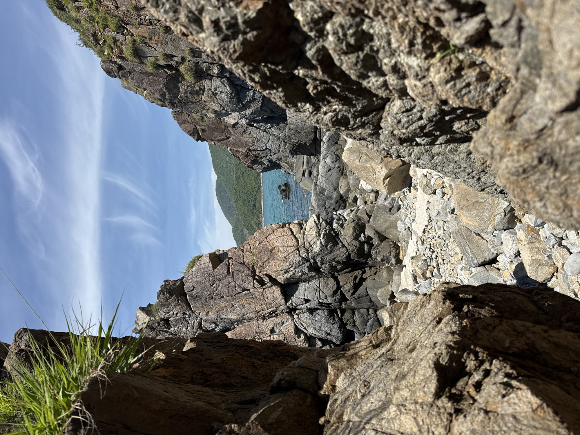 This is a photograph taken on a sunny day, looking through a rocky passage towards a body of water and a distant, tree-covered hill. The foreground is dominated by large, rough-textured rocks in shades of brown and gray, with some sparse green grass growing on the left side. The passage opens up to reveal a narrow strip of rocky beach and then the turquoise ocean. The water appears calm, with a few small rocks visible near the shore. In the background, a verdant hill rises from the water, its slopes covered in dense green vegetation. The sky is a clear blue with wispy white clouds. The scene appears to be a natural coastal environment, likely a beach or cove, located in the city of Vĩnh Lương, Vietnam.