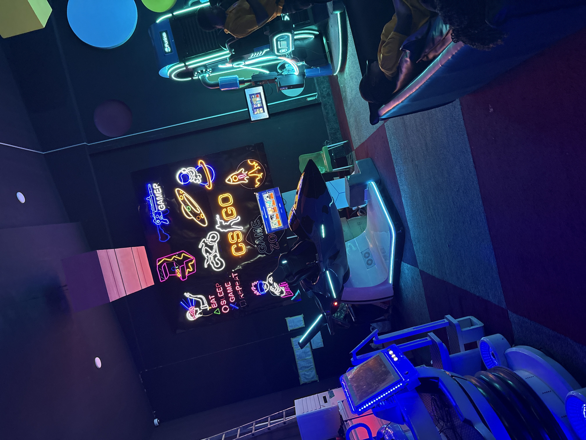 The image depicts an indoor entertainment area with gaming setups, including racing simulator pods and neon signs related to gaming. One sign reads "GAMER" vertically and shows various game-related symbols. Another sign has the phrase "EAT SLEEP GAME REPEAT". There are colored circular decorations on the ceiling, including blue, green, and pink. The specific location in Abuja, Nigeria, is not identifiable from the image.