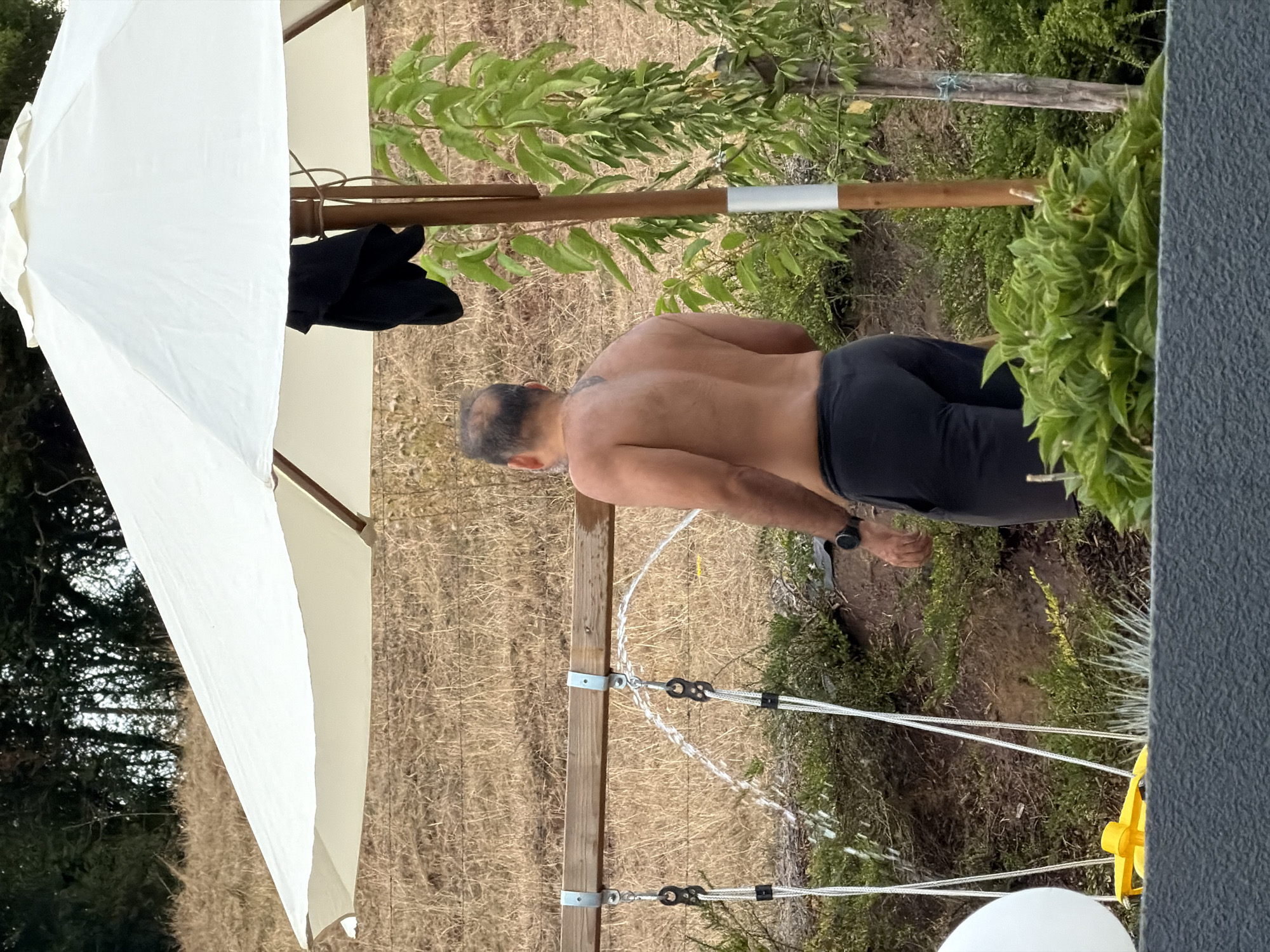 A man is standing shirtless and facing away from the camera, under a large white umbrella. He is wearing dark pants and a watch on his left wrist. Water is spraying from two spouts mounted on a wooden beam behind him. The environment appears to be outdoors with dry, brown grass in the background and green foliage in the foreground. The man has a tattoo on his upper back. A black garment is draped over the umbrella pole. A portion of a white sphere and a yellow object are visible in the lower left corner of the image. The scene is likely a backyard or garden area.