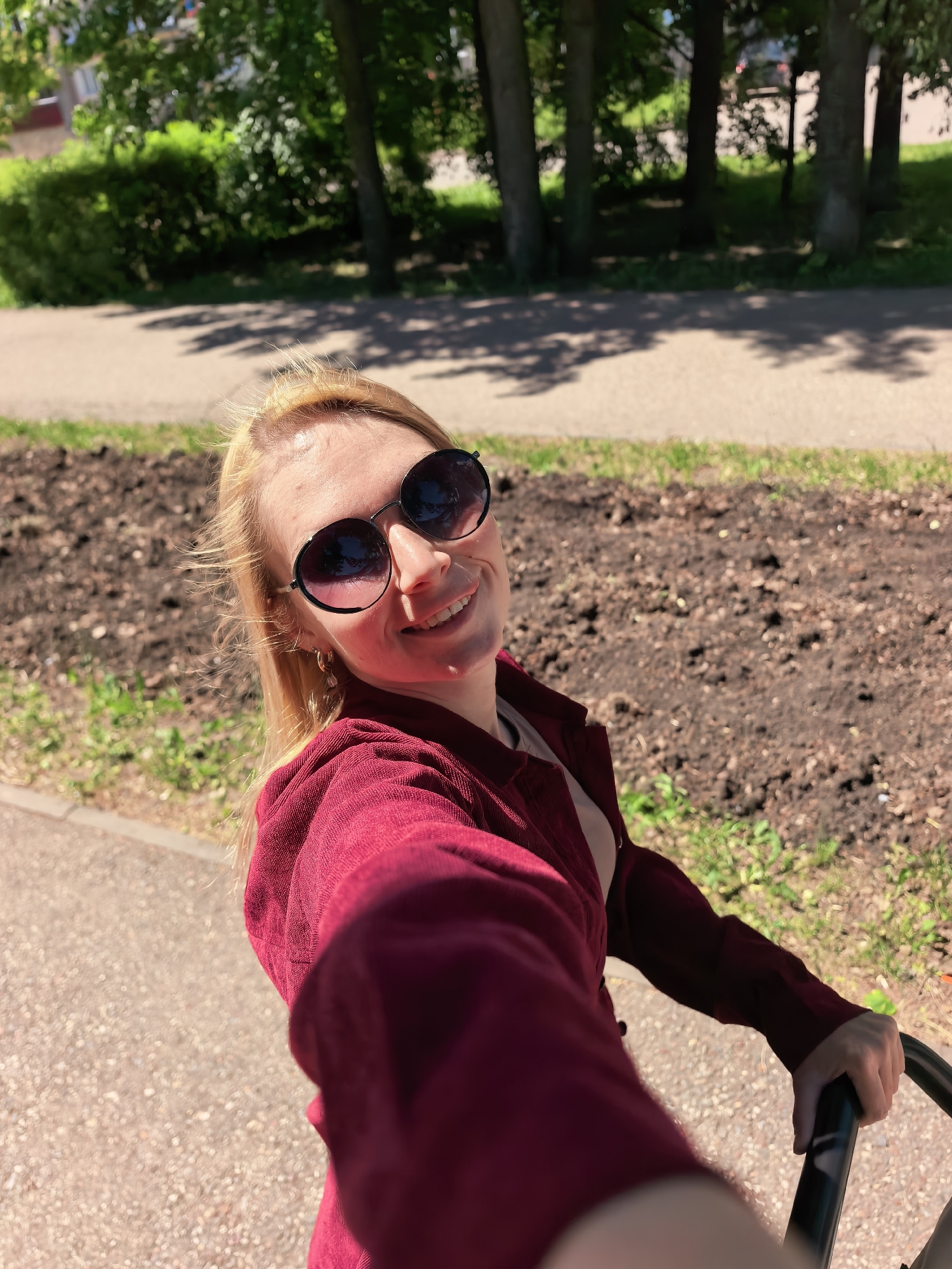 A blonde woman with sunglasses smiles at the camera in a selfie. She wears a maroon jacket. She is holding onto a black stroller frame. Behind her is a dirt patch with some grass. There is a sidewalk and trees in the background. The location is outdoors, likely in Sterlitamak, Russia.