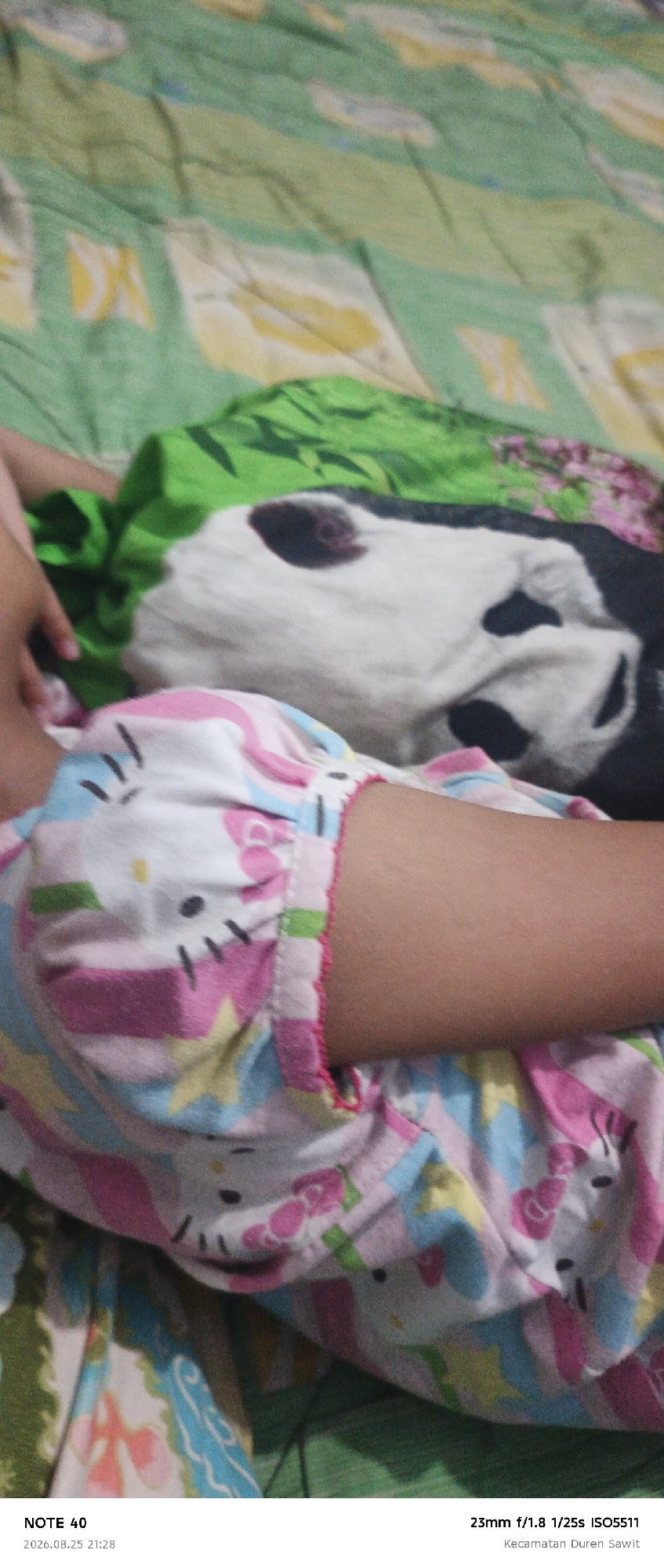 The image displays a close-up view of a child's arm and shoulder wearing pajamas with a pink and blue Hello Kitty pattern. The background consists of a green blanket with a colorful pattern, possibly a bedspread, and a portion of what appears to be a pillow or stuffed animal with black and white markings resembling a dog or panda. The lighting is dim, suggesting an indoor setting. The text at the bottom indicates the image was captured on a "NOTE 40" device on August 25, 2026, at 21:28, with camera settings of 23mm f/1.8 1/25s ISO5511, and mentions "Kecamatan Duren Sawit," which is a subdistrict in East Jakarta, Indonesia.