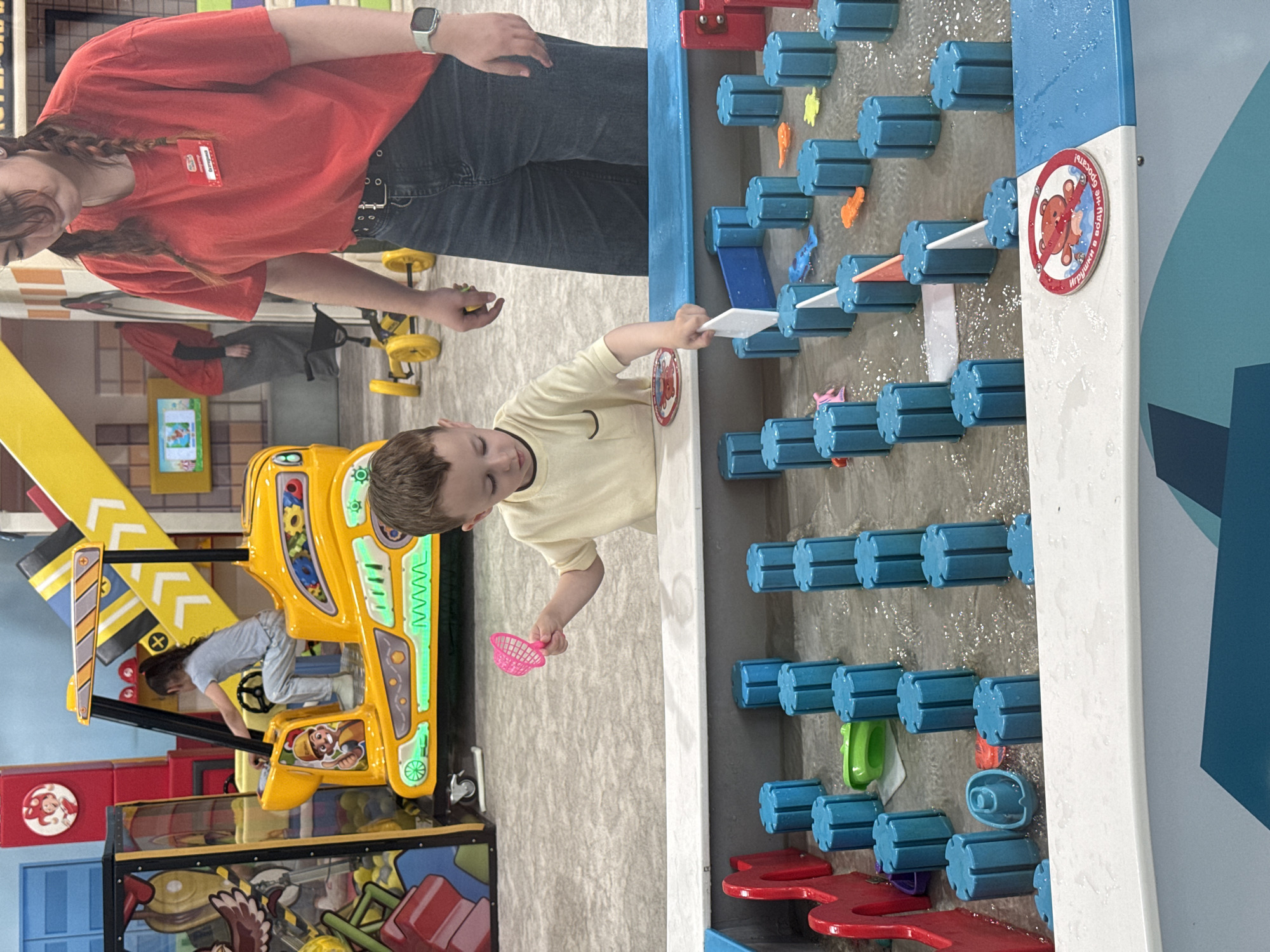 A child and an adult are at a water play table in an indoor play area. The child, wearing a light-colored shirt, is holding a white object and a pink net, and is focused on the water table. The adult, wearing a red shirt and blue jeans, stands beside the child. The water table features blue and red plastic components. A sign with a red teddy bear is visible on the side of the table. In the background are various other play structures, including a yellow crane-like structure and a climbing area.
The city of Vladivostok, Russia is not discernible from the given image.