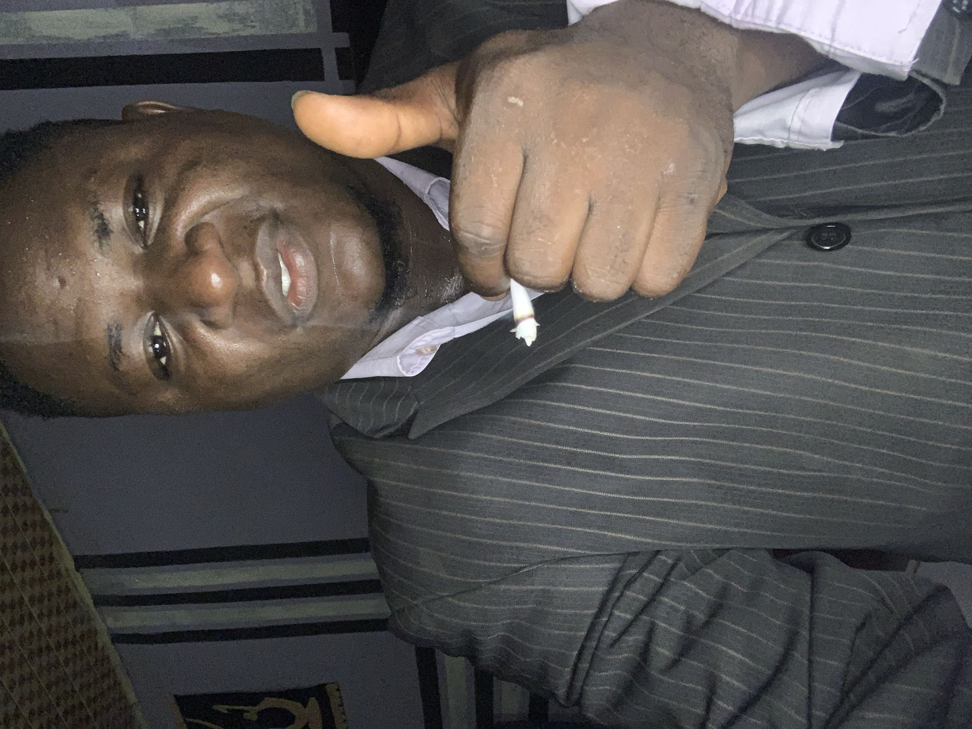 A man in a pinstripe suit is holding a lit cigarette between his fingers. The location is identified as Akwanga, Nigeria.