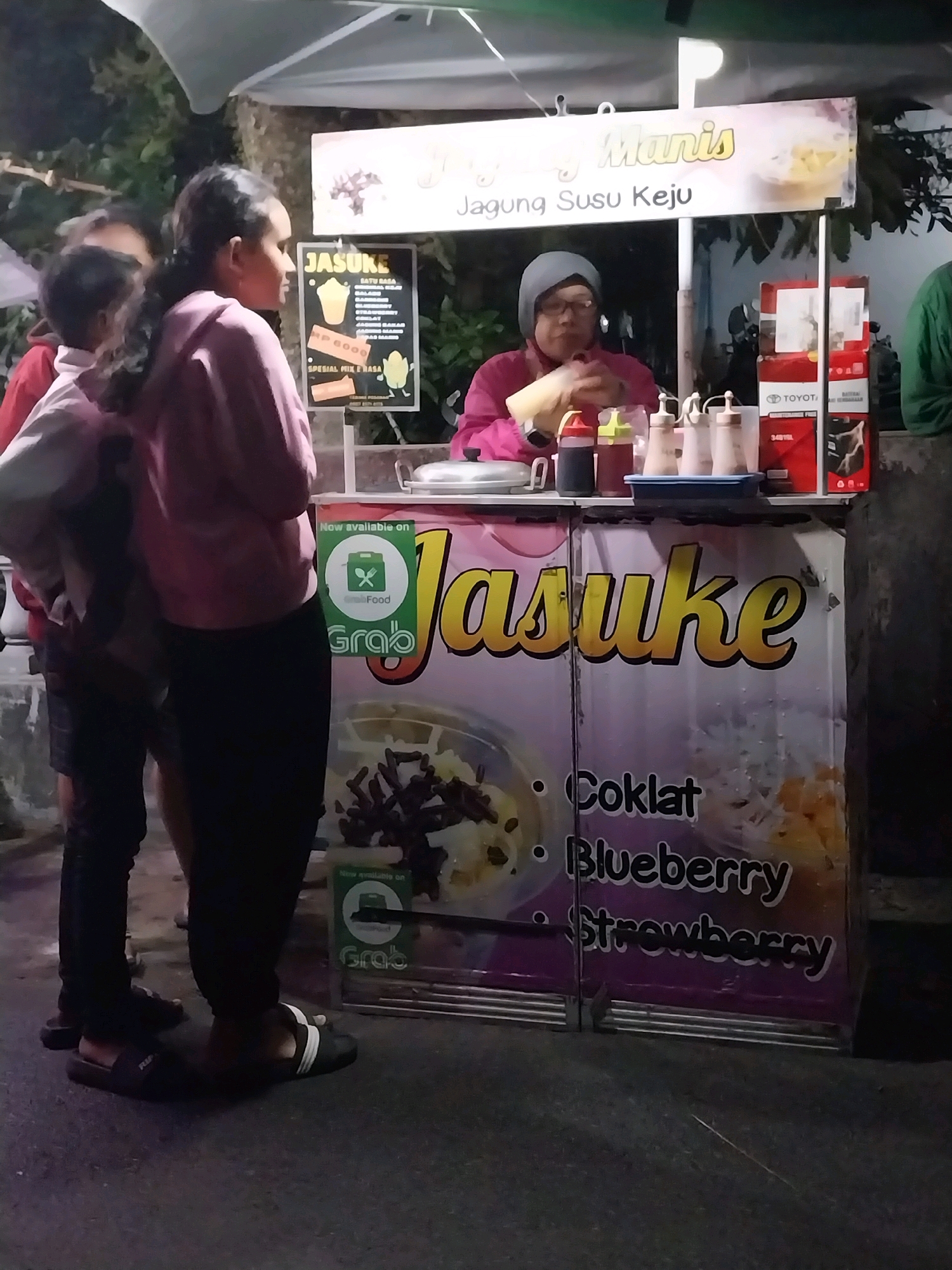 In the city of Nglegok, Indonesia, an outdoor food stall is in operation, likely at night given the ambient lighting. A woman, wearing a headscarf and glasses, is behind the counter, preparing or serving food. She has on a pink jacket. In front of the counter, two young individuals, a child and an older girl, are visible from the chest down. The girl is wearing a pink hooded sweatshirt and black pants, and the child is wearing a red jacket and black shorts. The stall's signage prominently features the word "JASUKE" in large yellow letters, accompanied by text in Indonesian that translates to "Sweet Corn Milk Cheese." Below this, a menu lists available flavors such as "Coklat" (Chocolate), "Blueberry," and "Strawberry." A "GrabFood" logo is also displayed, indicating the availability of delivery services. Several condiment bottles and containers are arranged on the counter. The overall scene suggests a casual food vending operation in a public space.