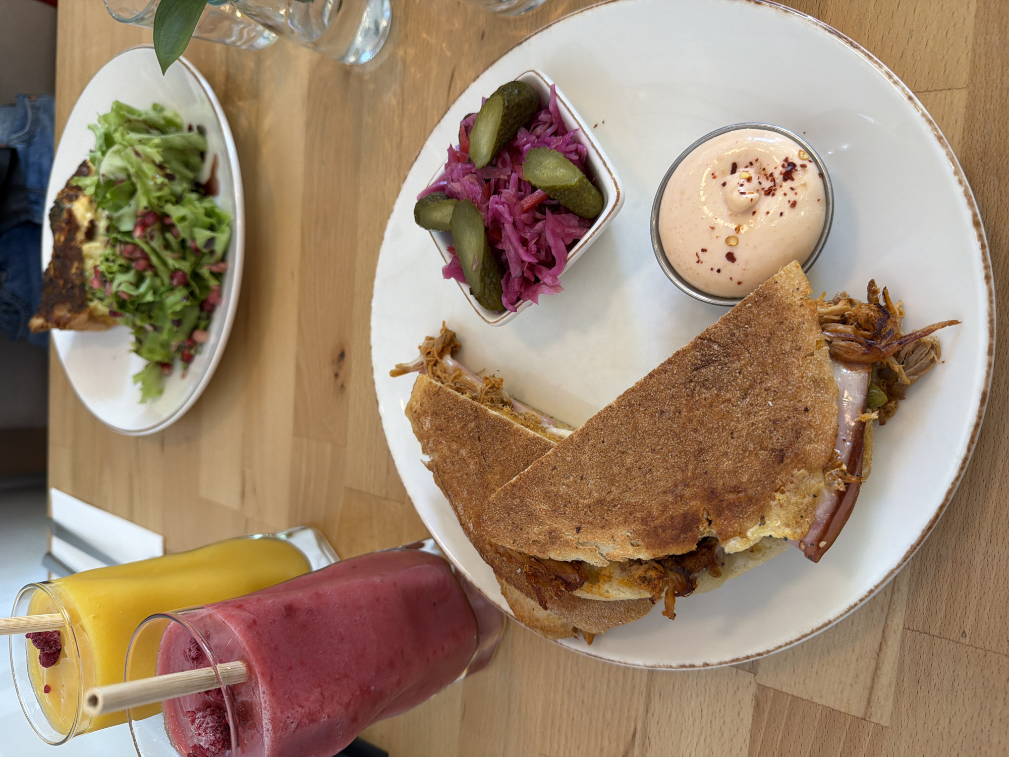 A high-angle shot shows a table setting with various food items and beverages. A white plate dominates the center of the frame, containing a sliced sandwich with a filling that appears to be pulled pork, a small white square bowl filled with pickled red cabbage and sliced pickles, and a small metal container filled with a sauce garnished with red pepper flakes. A second plate with a salad (likely containing pomegranate seeds and lettuce) and what appears to be a quiche is visible to the left of the main plate. In the foreground are two glasses, one filled with a yellow liquid and the other with a pink/red liquid, both with wooden sticks inside. The table is made of light wood. The location is likely in Sofia, Bulgaria, based on the style of the cafe.
