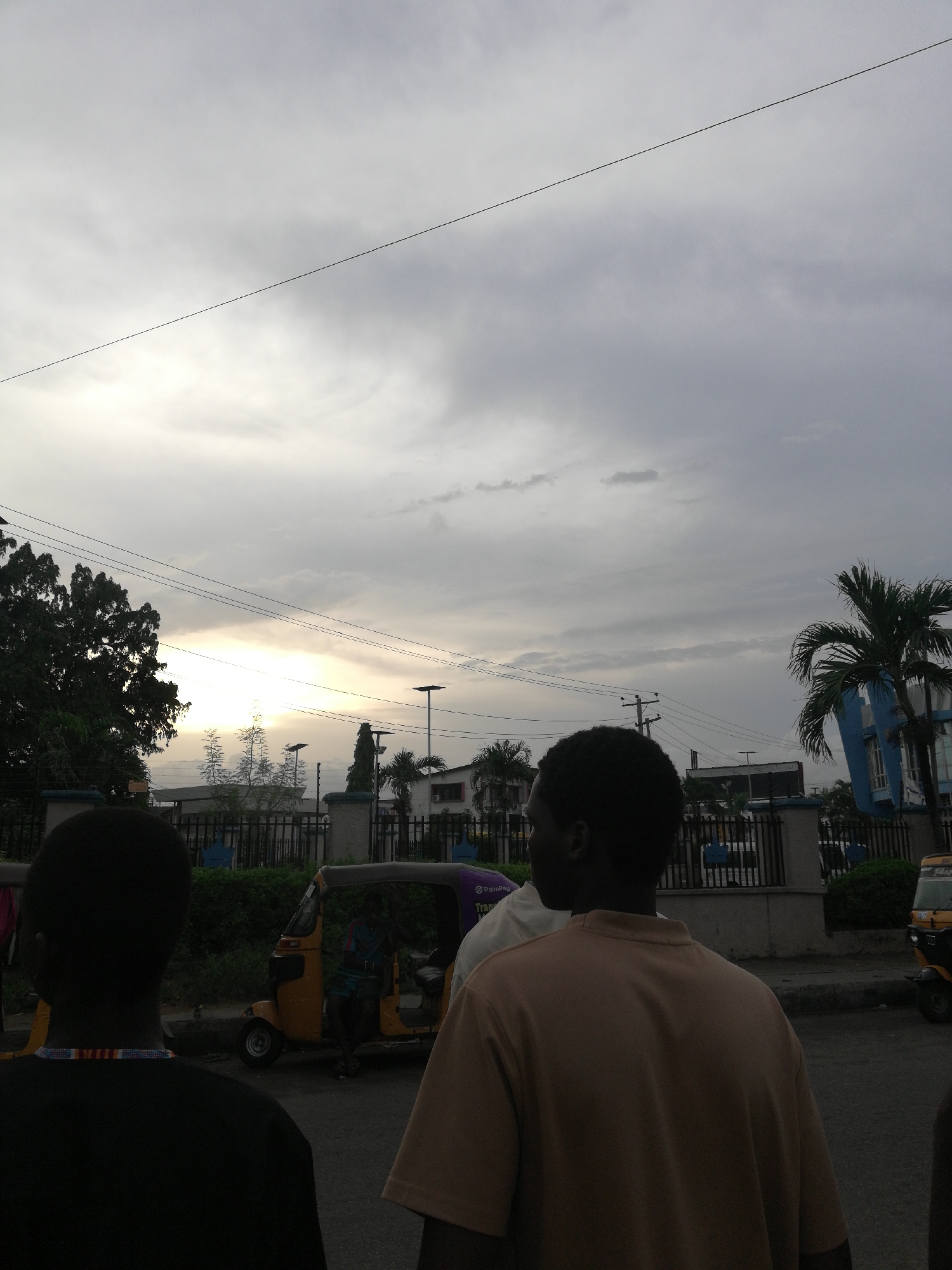 The image shows a street scene in Lagos, Nigeria under an overcast sky. Three people are in the foreground, with their backs to the camera. One person wears a black shirt with a colorful beaded necklace, while another wears a tan t-shirt. In the background, there is a line of auto rickshaws (tuk-tuks), one of which has a passenger. Behind them, there is a low fence with vegetation, and buildings are visible in the distance, including a blue and white structure with a palm tree nearby. Streetlights and power lines are visible in the sky. The overall atmosphere suggests early morning or late afternoon due to the muted lighting and cloudy sky.