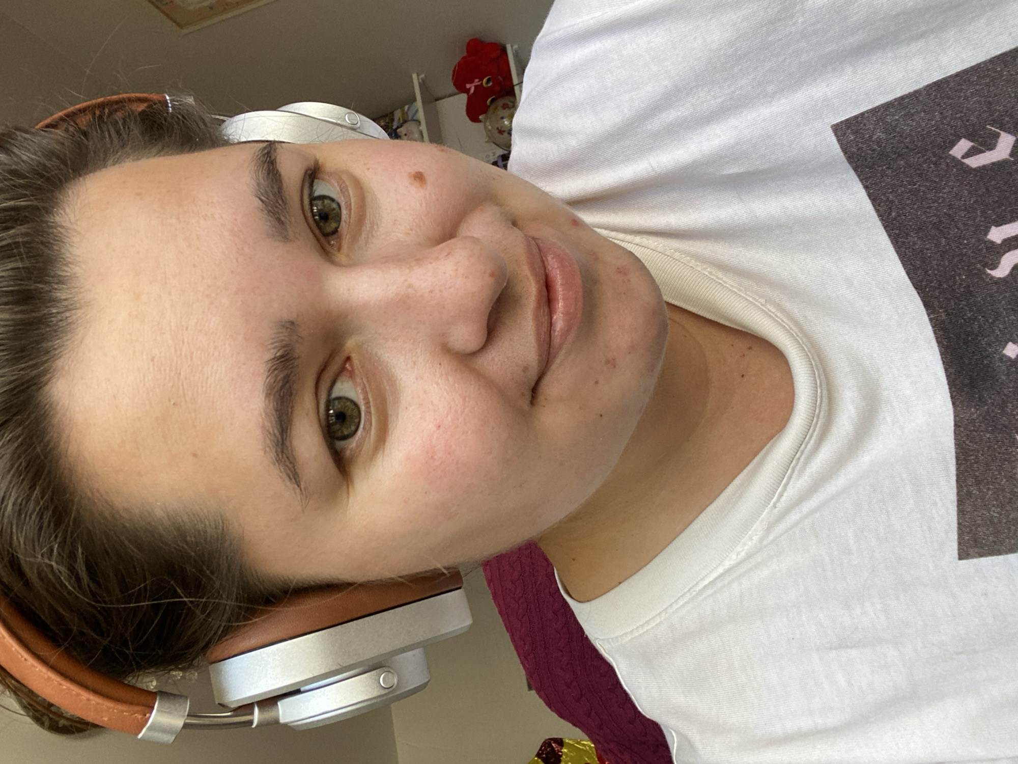 A close-up, overhead, medium shot shows a young woman with brown hair pulled back and wearing silver and tan headphones. She has fair skin, green eyes, and a light smudge of pink blush on her cheeks. Her head is tilted slightly to her right, and she is looking at the camera with a gentle smile. She is wearing a white t-shirt. In the background, a shelf with some stuffed animals and a framed picture can be seen. The woman's head and shoulders are the main focus of the image, and the background is blurred. The lighting appears to be natural, coming from the right side of the image. The scene appears to be indoors, possibly in a bedroom or living room.