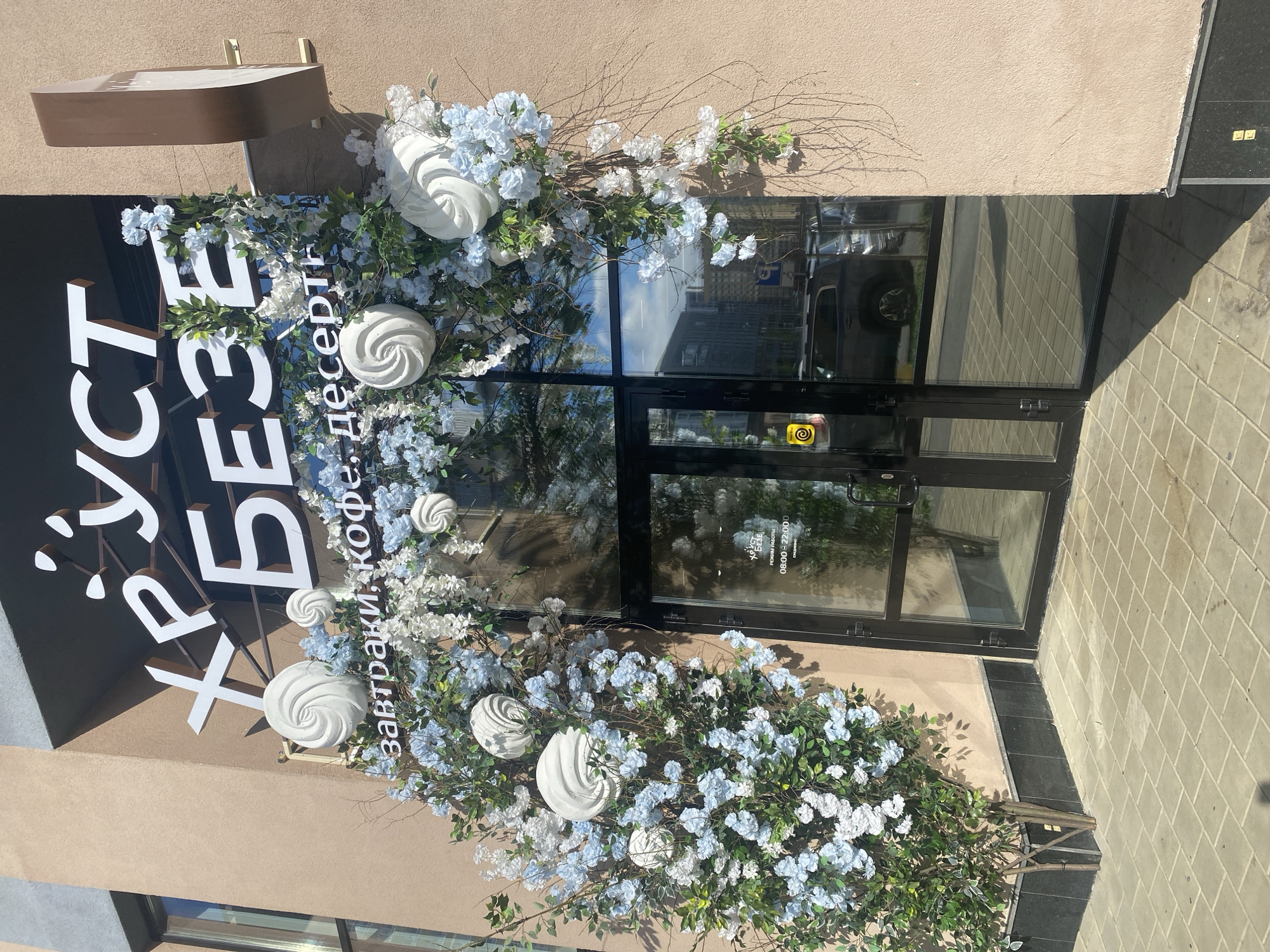 This is an exterior shot of a business in Yekaterinburg, Russia. The building is adorned with light blue and white flowers, and meringue-shaped decorations. Signage indicates the business name is "Хруст Беззе" (Khrust Beze) or "Crunchy Meringue." Another sign states "завтраки, кофе, десерт," which translates to "breakfast, coffee, dessert." The shop hours are shown as 08:00 - 22:00. The business has glass doors and appears to be located on a sidewalk. The photo is taken during daylight hours.