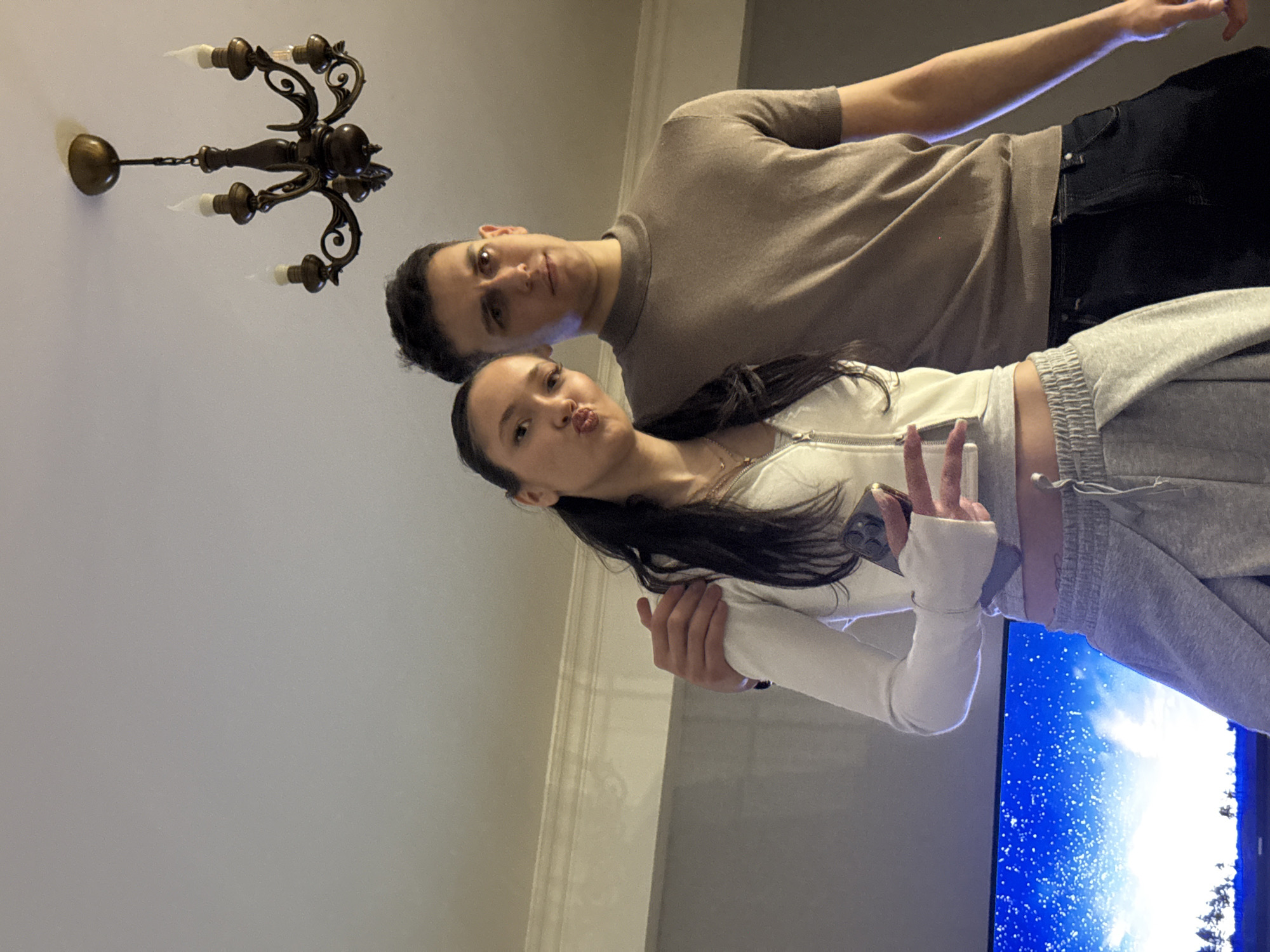 A couple poses indoors for a photo. The man is behind the woman, with his arm around her. He wears a brown t-shirt and dark pants. The woman wears a white, cropped zip-up jacket and gray sweatpants. She puckers her lips and holds up two fingers in a peace sign. A chandelier is visible on the ceiling. A television screen displays a scene of a sunny sky. The location is likely a private residence in Yerevan, Armenia.