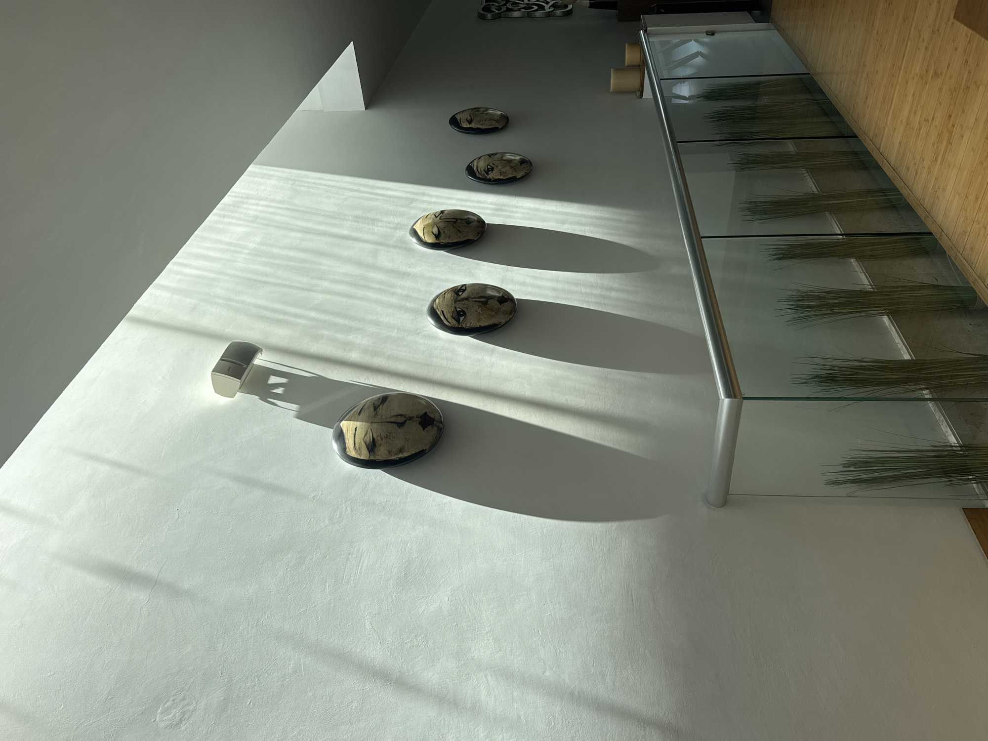 The photograph depicts an interior architectural scene, likely inside a building in San Bernardino Tlaxcalancingo, Mexico. The focus is on a light-colored wall onto which five circular objects with dark edges are mounted in a vertical row. These objects appear to have an image or design on their surface.
Light falls across the wall, creating strong linear shadows that intersect the objects and the wall’s surface. To the right of the objects is a structure with multiple horizontal glass shelves containing straw-like objects. The structure is supported by metal posts.
A small, metallic object is affixed to the wall just above the lowest circular object. The wall surface is textured with some subtle imperfections. The overall impression is of a minimalist and artful interior design.