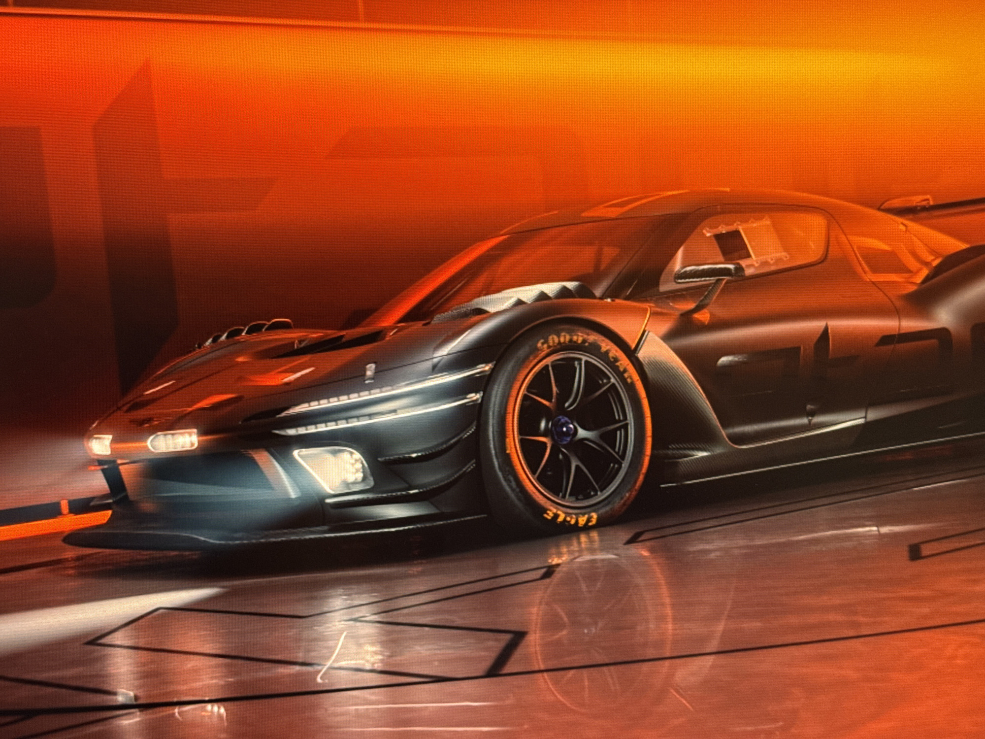 The image shows a dark gray, futuristic-looking race car with orange accents on the tires. The car is positioned at an angle, with its front left tire prominently featured. The car's body appears to be made of carbon fiber and has sharp, aerodynamic lines. It has bright LED headlights. The background is a vibrant orange, with a textured surface that suggests a large screen or backdrop. The floor beneath the car is reflective, showing a distorted reflection of the vehicle. The overall lighting is dramatic, with strong orange and red hues dominating the scene. There is no indication of the city of Baku, Azerbaijan, in the image. The scene appears to be a studio or exhibition setting for showcasing the car. There are no people or any other objects or activity depicted.