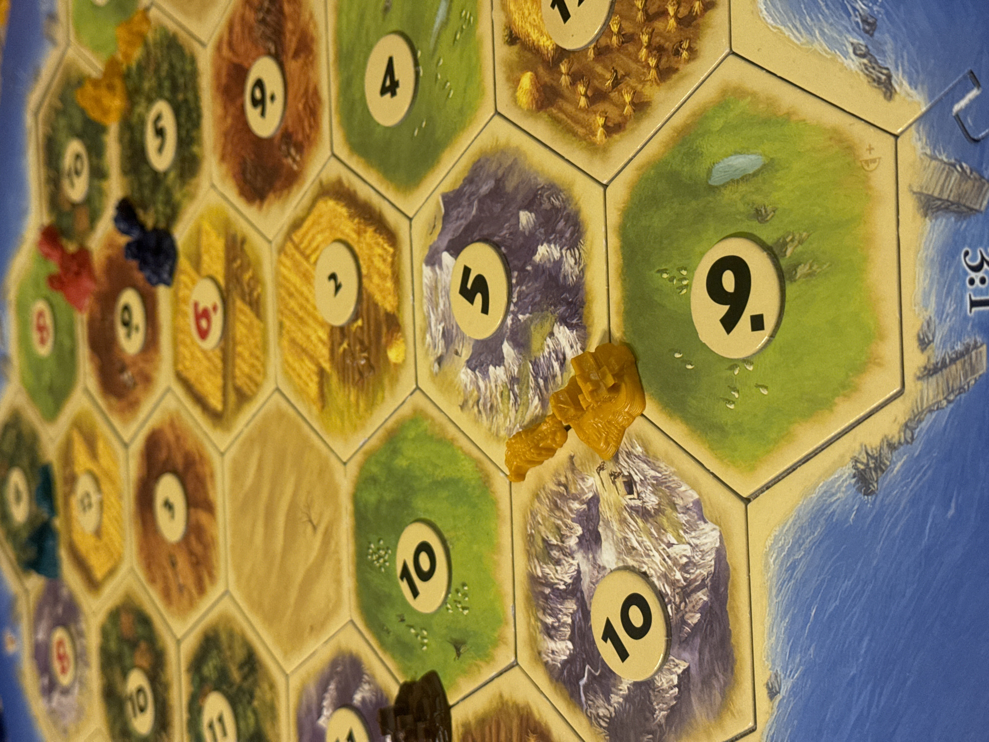 The image shows a section of a Catan board game. The board consists of hexagonal tiles representing different types of terrain: fields of wheat, hills of brick, mountains of ore, forests of lumber, pastures of sheep, and a desert. Each tile has a numbered token, ranging from 2 to 12, which determines when players receive resources from that tile. Small plastic pieces, representing settlements, roads, and cities, are placed along the edges of the tiles. In the visible section of the board, there are several colored settlements. The environment is a domestic setting, likely indoors, where the game is being played. There is no indication of Krasnoyarsk, Russia.