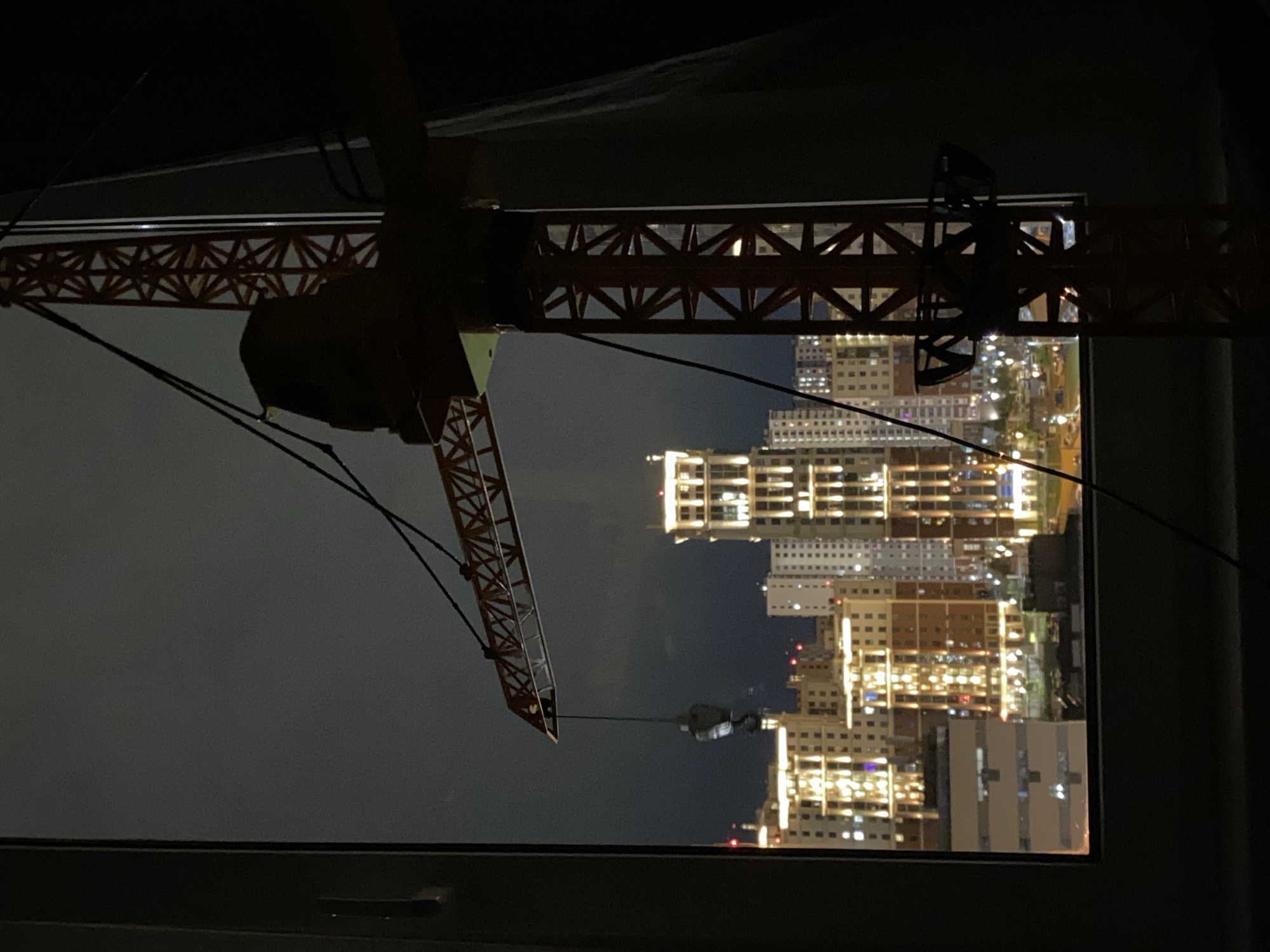The image shows a view from a window at night, with a construction crane prominently visible in the foreground. Through the window, there is a cityscape with multiple illuminated buildings. The style of the architecture and the overall appearance of the buildings suggest this may be Saint Petersburg, Russia. However, definitive confirmation is not possible from this image alone without additional context or information.