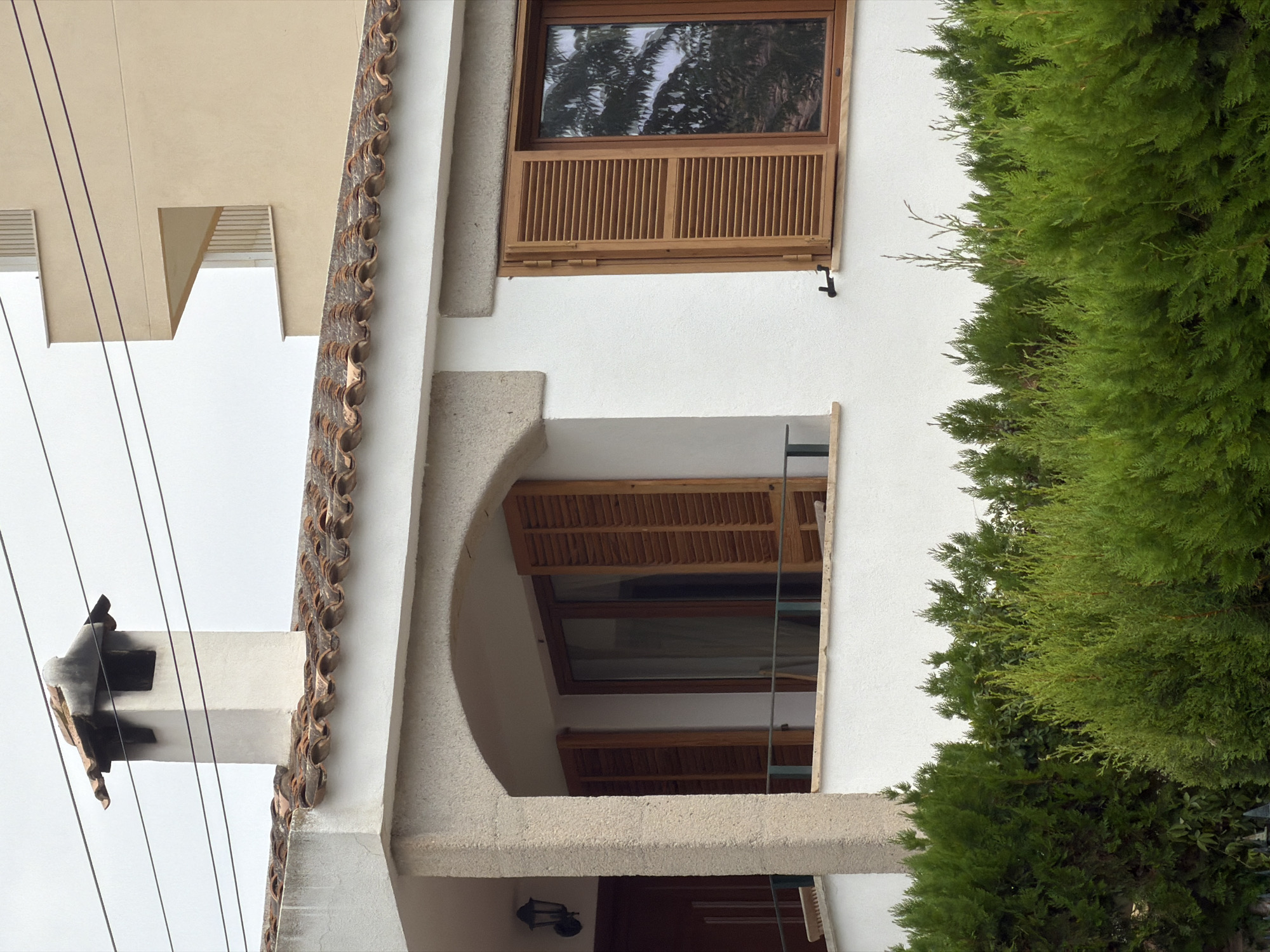 The image shows the exterior of a multi-story building, likely residential, in Palmanova, Spain. The foreground is dominated by a dense hedge of green evergreen shrubs. Behind the hedge, the lower facade of the building is visible, painted white and featuring a white stone column supporting an arched overhang. To the left of the column, a dark-colored door and an outdoor lamp are partially visible.
Further to the right, there is a window with wooden shutters that are partially open, revealing a view of curtains inside. The window frame and shutters are made of wood. Above this, the tiled roof of the building is visible, made of reddish-brown, curved tiles. A white chimney with a dark cap extends above the roofline, with electrical wires running horizontally behind it.
In the background, another building is visible, constructed with light beige stucco and featuring balconies with white railings. The sky is overcast. The architectural style suggests a Mediterranean setting.