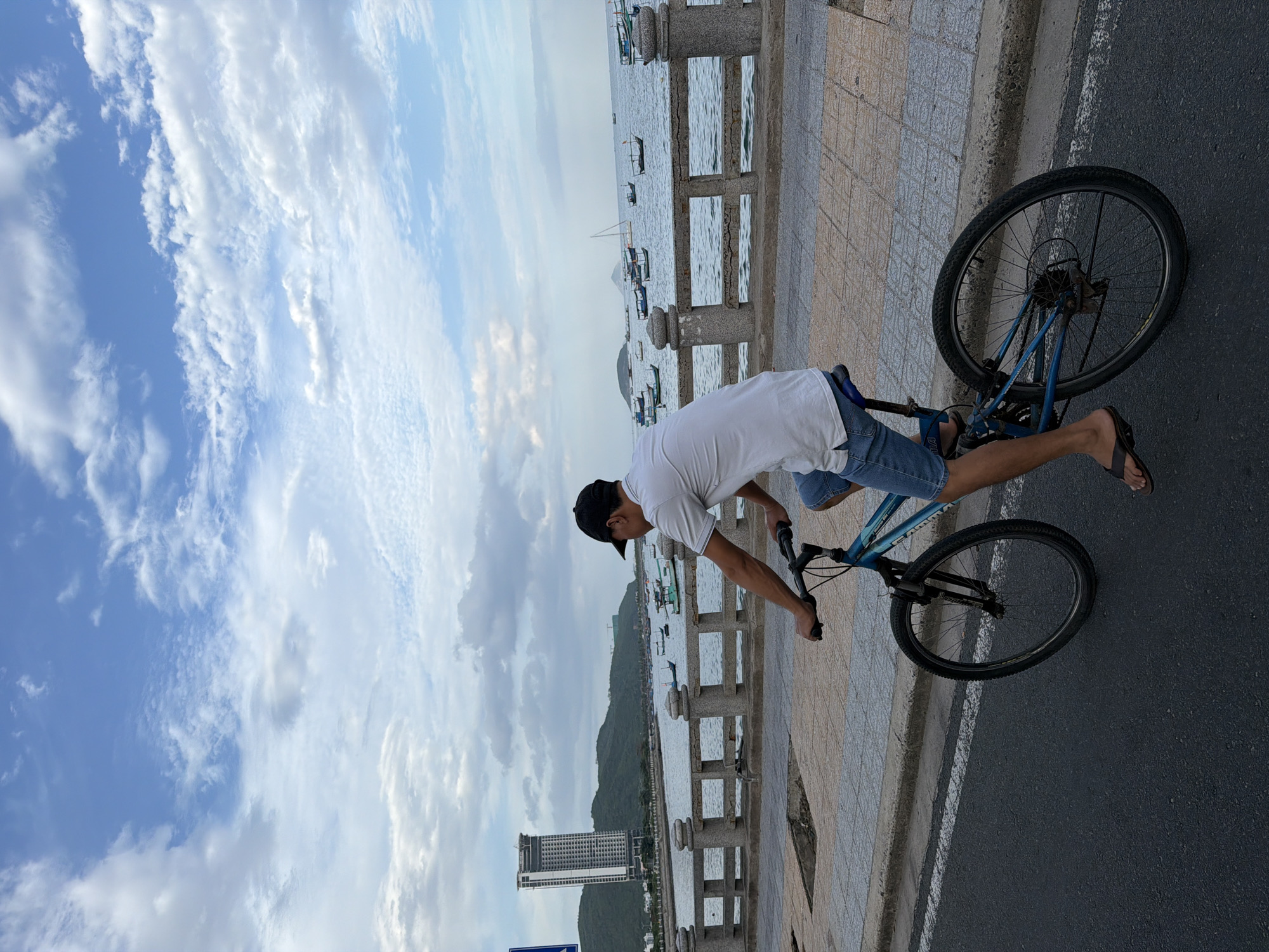 A man wearing a white polo shirt, blue denim shorts, and a black baseball cap is riding a blue bicycle along a paved road next to a body of water. The man is positioned in the right-middle of the frame, facing away from the viewer and towards the water. His left arm is extended, holding the handlebar, while his right arm is also on the handlebar. He is wearing flip-flops. The bicycle has black tires and a blue frame.
The road has a white line marking the edge. To the left of the road is a pedestrian walkway made of interlocking square tiles. A grey stone railing with rounded pillars separates the walkway from the water. Beyond the railing, a calm body of water is visible, dotted with numerous small boats, some with sails, and a larger white sailboat. In the distance, green hills rise up, and on one of the hills, a tall, modern, white-and-grey building with many windows stands out. The sky above is blue with scattered white and grey clouds. The overall scene suggests a coastal urban environment. The location is likely along the coast of Nha Trang, Vietnam, given the architecture and the presence of many boats.