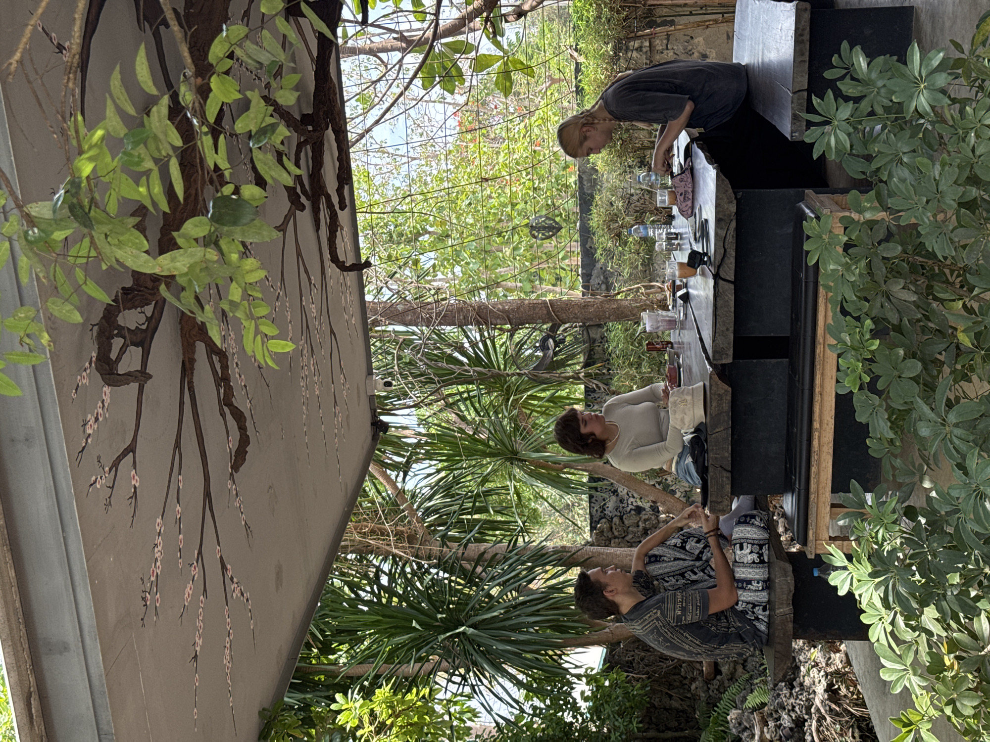 The image depicts an outdoor scene in Ban Tom, Thailand, featuring three people engaged in different activities. One person with short hair is standing behind a counter, possibly preparing drinks or food. Two other individuals are seated at the counter, facing each other. One is dressed in a patterned outfit, while the other is wearing a light-colored shirt. The counter is set with various items, including glasses and bottles. The setting appears to be a restaurant or bar, surrounded by lush greenery, including trees and plants. A structure with a tree branch design is visible in the background.