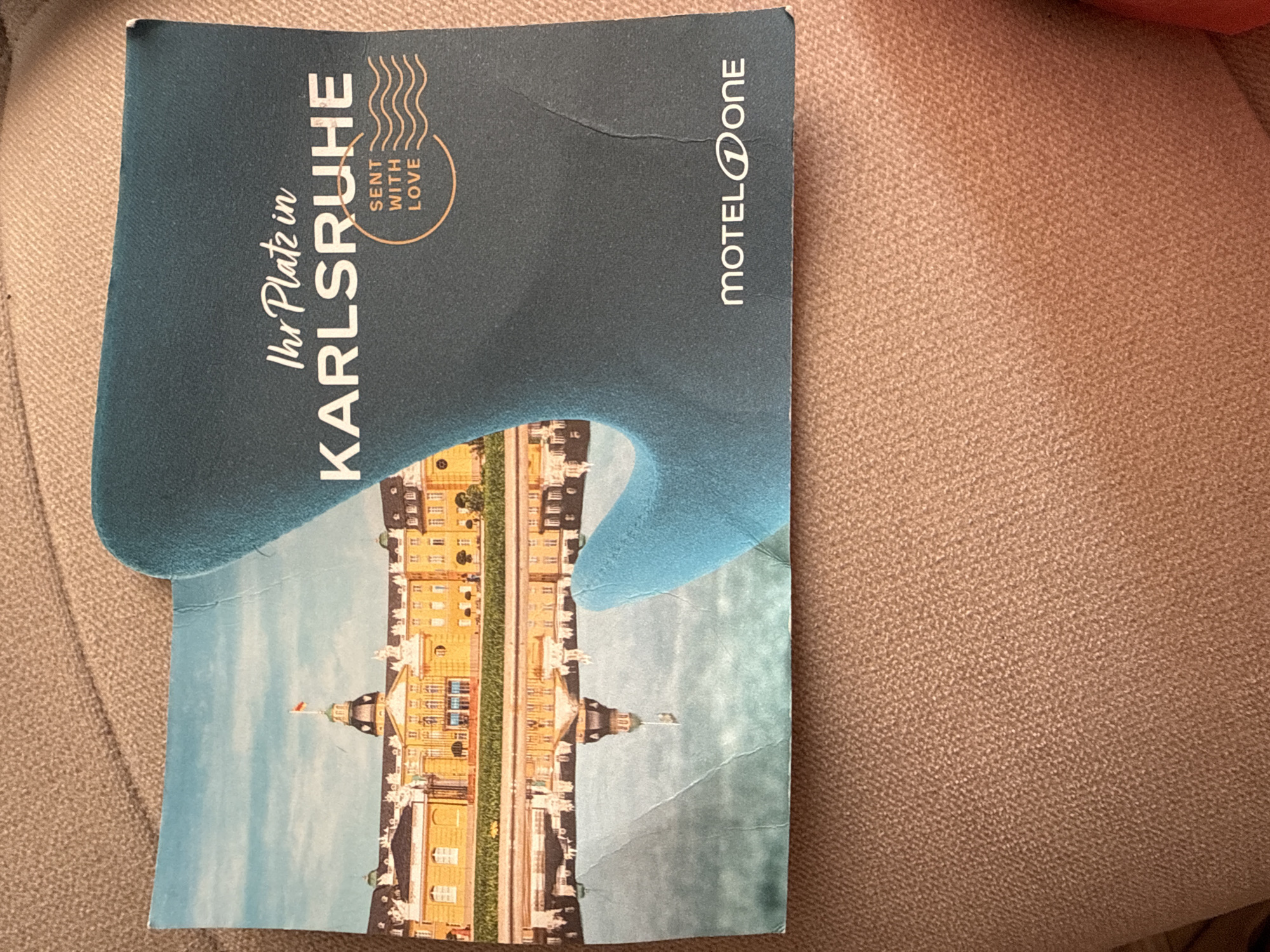 A slightly creased and worn, vertically oriented postcard or folded card rests on a light brown textured fabric surface. The top portion of the card is a dark blue-green, featuring white text and a distinctive, gentle curvilinear shape along its lower edge, bordering the main illustration.
The lower two-thirds of the card display a vibrant illustration or photograph of a grand, yellow-toned European palace or castle with a central dome or tower and numerous windows. Below the building, a detailed reflection is visible, creating a mirrored image that suggests either a body of water in the foreground or a stylized design. The sky above the palace is a clear light blue, indicating daytime.
Prominent white text on the dark blue-green section reads "Ihr Platz in" in a handwritten-style script, followed by "KARLSRUHE" in large, bold capital letters. To the right of "KARLSRUHE," a circular design resembling a postmark contains the phrase "SENT WITH LOVE," with three wavy lines above it. In the top right corner of the card, the brand name "MOTEL ONE" is visible, with a stylized 'O' in 'ONE'. The card exhibits minor creases and folds, particularly across the illustrated palace and its edges. The background fabric is out of focus, highlighting the card.