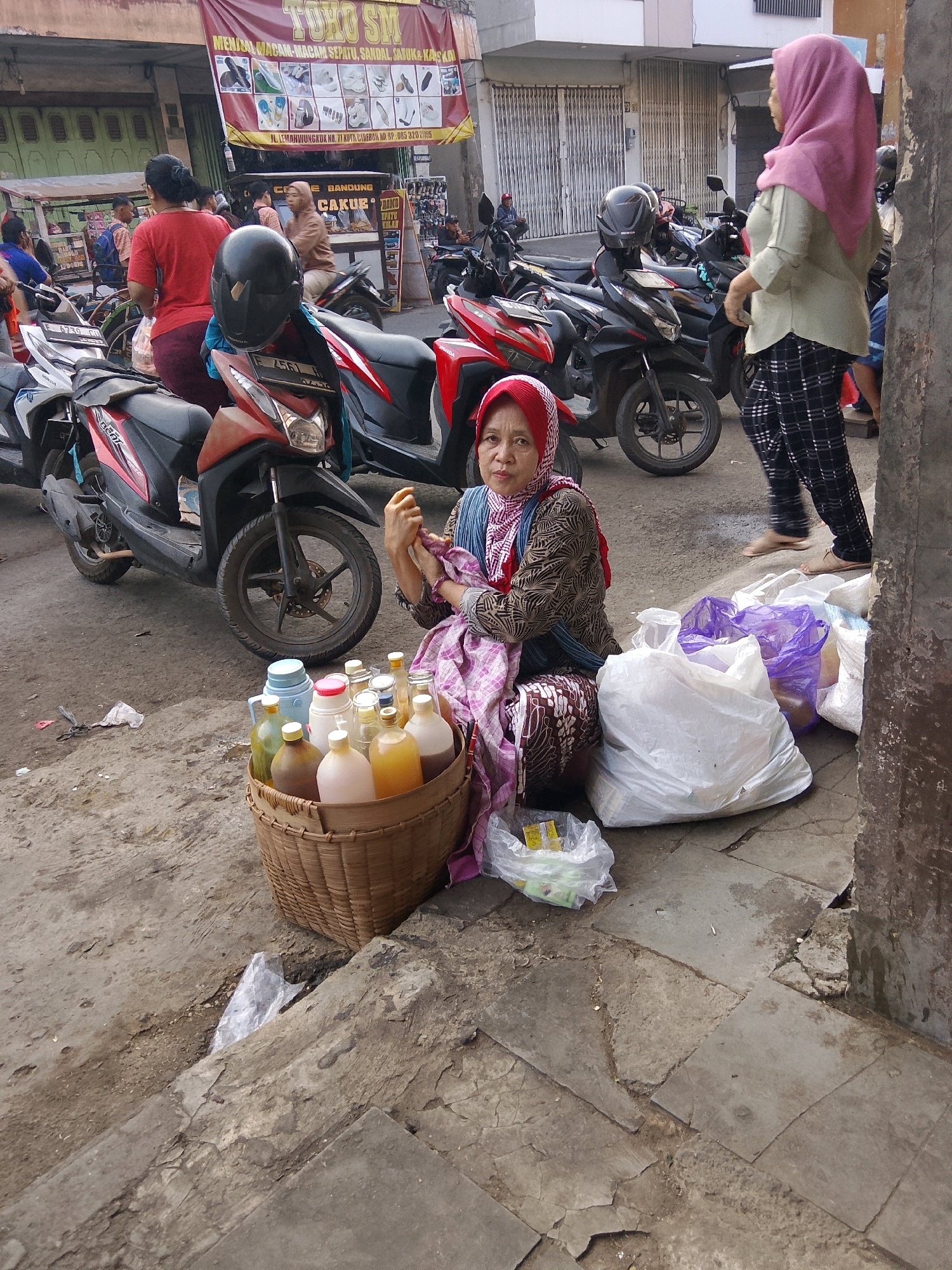 The image depicts a street scene in Cirebon, Indonesia, likely a marketplace or a busy thoroughfare. In the foreground, a woman wearing a red and white headscarf and a patterned long-sleeved shirt sits on the ground next to a woven basket filled with various bottles containing liquids of different colors. She is holding a small item in her hands and appears to be interacting with it. Beside her are several large plastic bags, some white and some purple. Behind her, a red and black motorbike is parked.
In the mid-ground, more motorbikes are visible, suggesting this is an area where vehicles are commonly parked or where traffic is present. A woman in a light green shirt and plaid pants walks away from the viewer towards the right of the frame. Further in the background, a banner is displayed overhead with text and images of footwear. There are buildings with metal shutters and a small food stall. Several other people are present in the background, some on motorbikes and others walking. The ground is paved with concrete and appears somewhat worn and uneven. The overall atmosphere suggests a bustling urban environment.