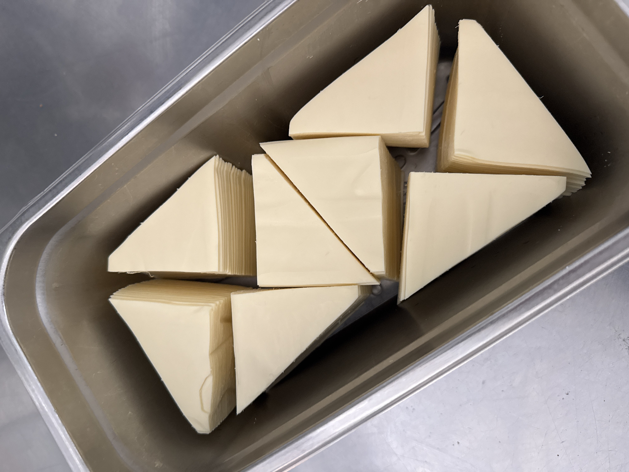 In an overhead shot of a stainless steel rectangular pan, several stacks of triangular slices of cheese are arranged. The cheese appears to be light yellow and is neatly stacked, with some stacks cut into half triangles. The pan is filled with these cheese slices, which are positioned at various angles within the container. The background is a metallic surface.
