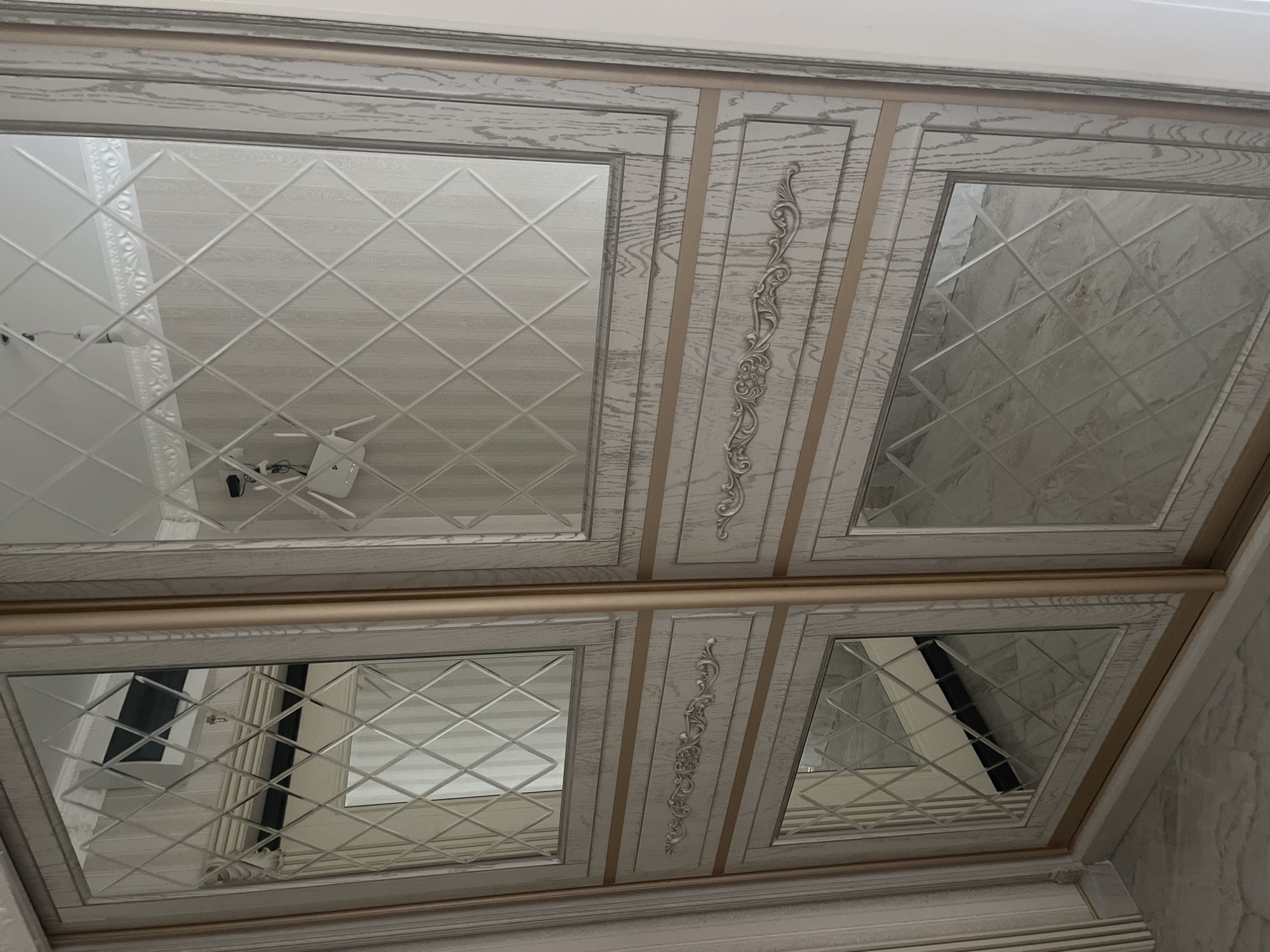 The image shows a close-up view of a wardrobe's sliding doors, featuring a combination of wood-effect panels and diamond-patterned mirrored sections. The wardrobe is light-colored with a visible wood grain texture and accented with thin, metallic gold trim. Each door is divided into three horizontal sections, with mirrored panels at the top and bottom, and a decorative panel in the middle. The central decorative panels have ornate, sculpted embellishments. The mirrored sections reflect the interior of the room, including a light-colored wall with vertical lines and a white ceiling with decorative molding. In the upper left mirrored section, a black rectangle, likely an air conditioner unit, is visible. In the upper right mirrored section, a white Wi-Fi router is mounted on the wall. The floor appears to be tiled with a marble-like pattern. This wardrobe is located in an interior space in the city of Kommunarka, Russia.