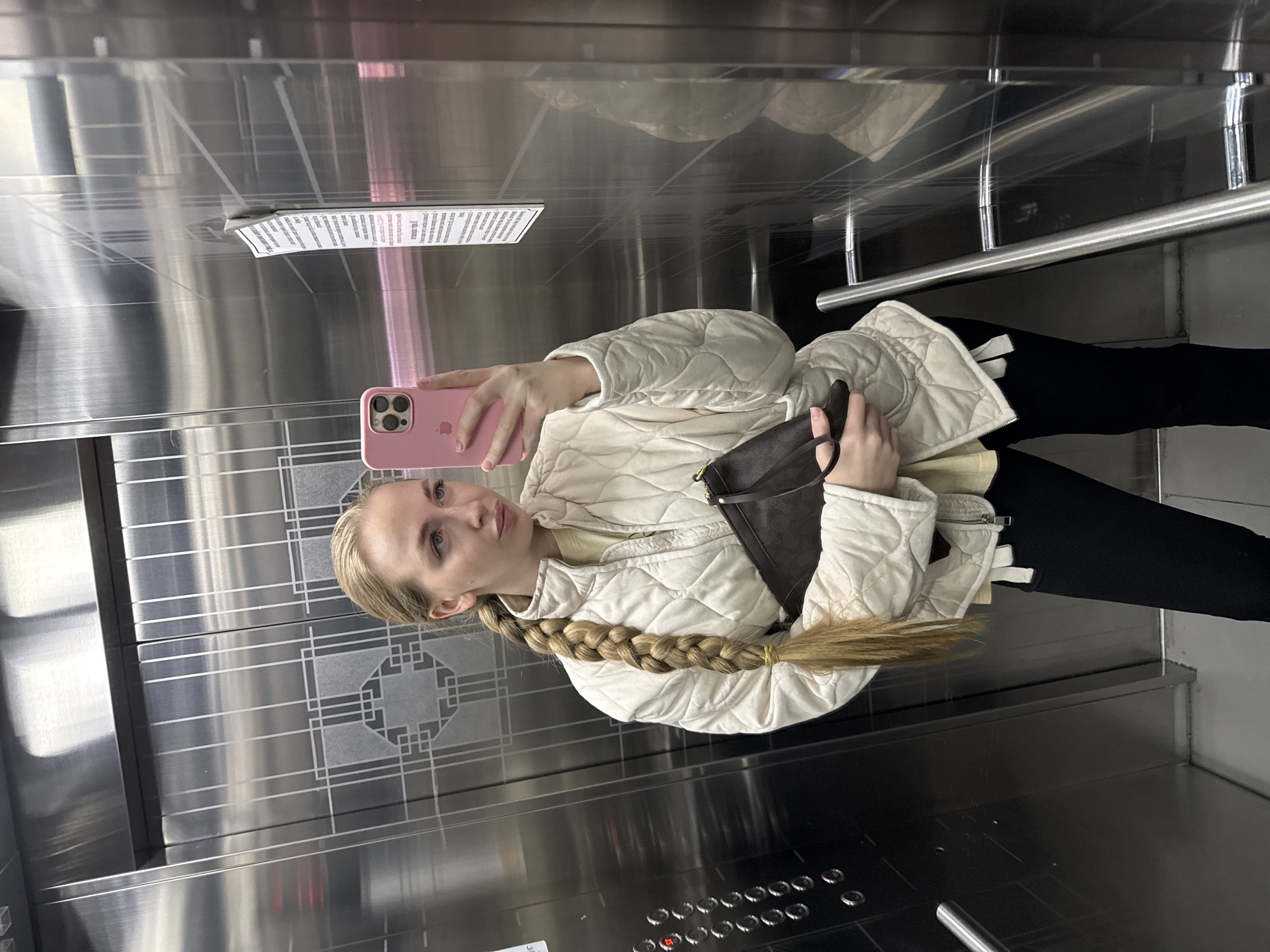 A young woman is captured in a mirror selfie taken inside an elevator. The image is rotated 90 degrees clockwise.
**Subjects:**
The primary subject is a woman with long, blonde hair styled in a thick braid that reaches below her waist. She is wearing a light cream or off-white quilted jacket over a light yellow top and black pants. She holds a pink smartphone (appears to be an iPhone with a triple camera system, with a faint Apple logo visible) in her left hand, which she uses to take the photo, and a dark brown clutch or small bag in her right hand. Her expression is neutral as she looks towards the camera reflected in the elevator wall.
**Scene and Setting:**
The setting is the interior of an elevator, characterized by highly reflective, likely brushed stainless steel walls. The walls feature patterned etchings, possibly decorative. The floor appears dark. A horizontal metal handrail is visible on one side. A control panel with multiple rows of circular metallic buttons is located below the woman's right arm. One button on the panel shows a red light, possibly indicating a current floor or selection.
**Activity:**
The woman is taking a mirror selfie, using the reflective surface of the elevator wall.
**Notable Details:**
The reflective nature of the elevator walls creates distorted reflections of the woman and the elevator interior. A piece of white paper, possibly a notice or information sheet, is attached to the upper part of one wall within a clear holder.
**Visible Text:**
Text is visible on the paper notice attached to the wall, but it is small, distant, and partially obscured by reflections, rendering it largely illegible. The Apple logo is faintly visible on the back of her pink phone.