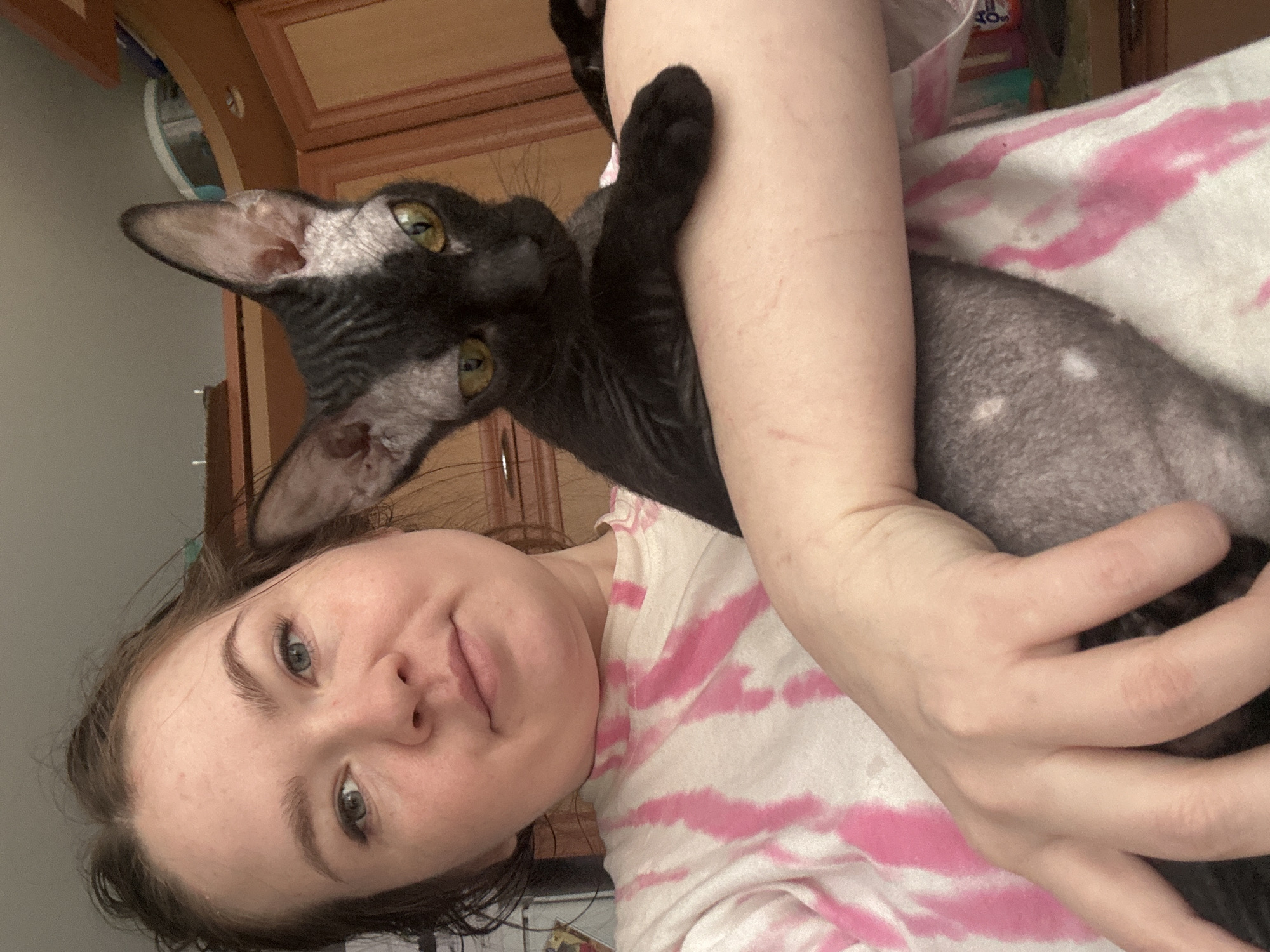 A woman is holding a black cat. The woman has fair skin and green eyes. She is wearing a white t-shirt with pink tie-dye. The cat has large ears and yellow eyes. The background is blurry and shows wooden cabinets and a white door. The location and city of Chaikovsky, Russia are not evident.