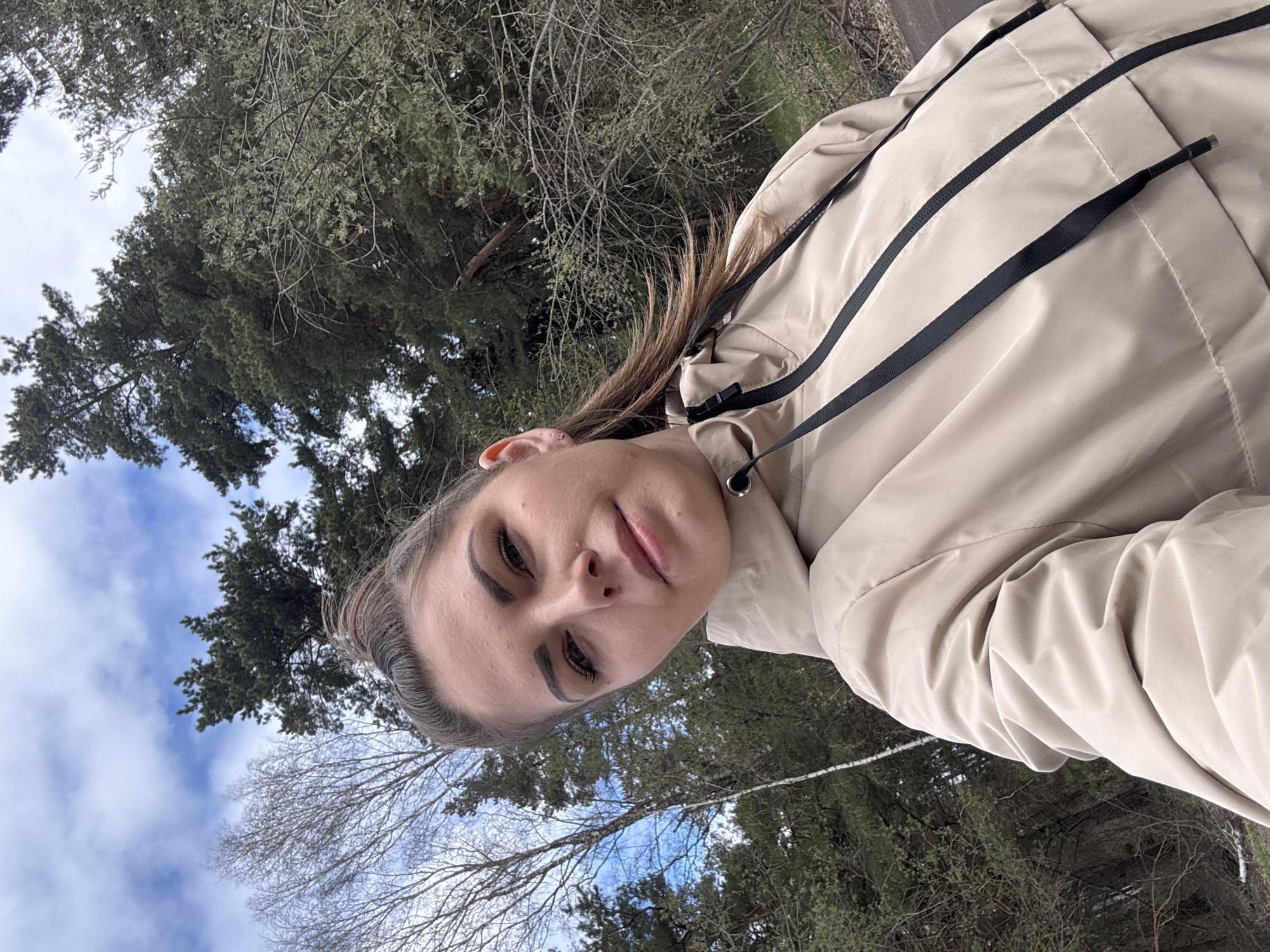 The image is a portrait-oriented outdoor selfie, presented sideways, depicting a young woman against a natural backdrop. When viewed upright, the woman is in the foreground, looking directly at the camera with a calm, neutral expression. She has medium-length brown hair, pulled back, and prominent dark eyebrows and eyelashes. She is wearing a light beige or khaki hooded jacket, featuring black drawstrings around the hood and collar.
The background reveals a natural, wooded environment, likely a forest or park. It includes a mix of dense evergreen coniferous trees with dark green foliage and deciduous trees with bare branches, suggesting either late winter or early spring. Above the trees, the sky is partly cloudy, showing patches of blue amidst white clouds. The natural lighting indicates it is daytime. The overall setting is a serene outdoor landscape, consistent with a location such as Раздолье, Russia. There is no visible text in the image.