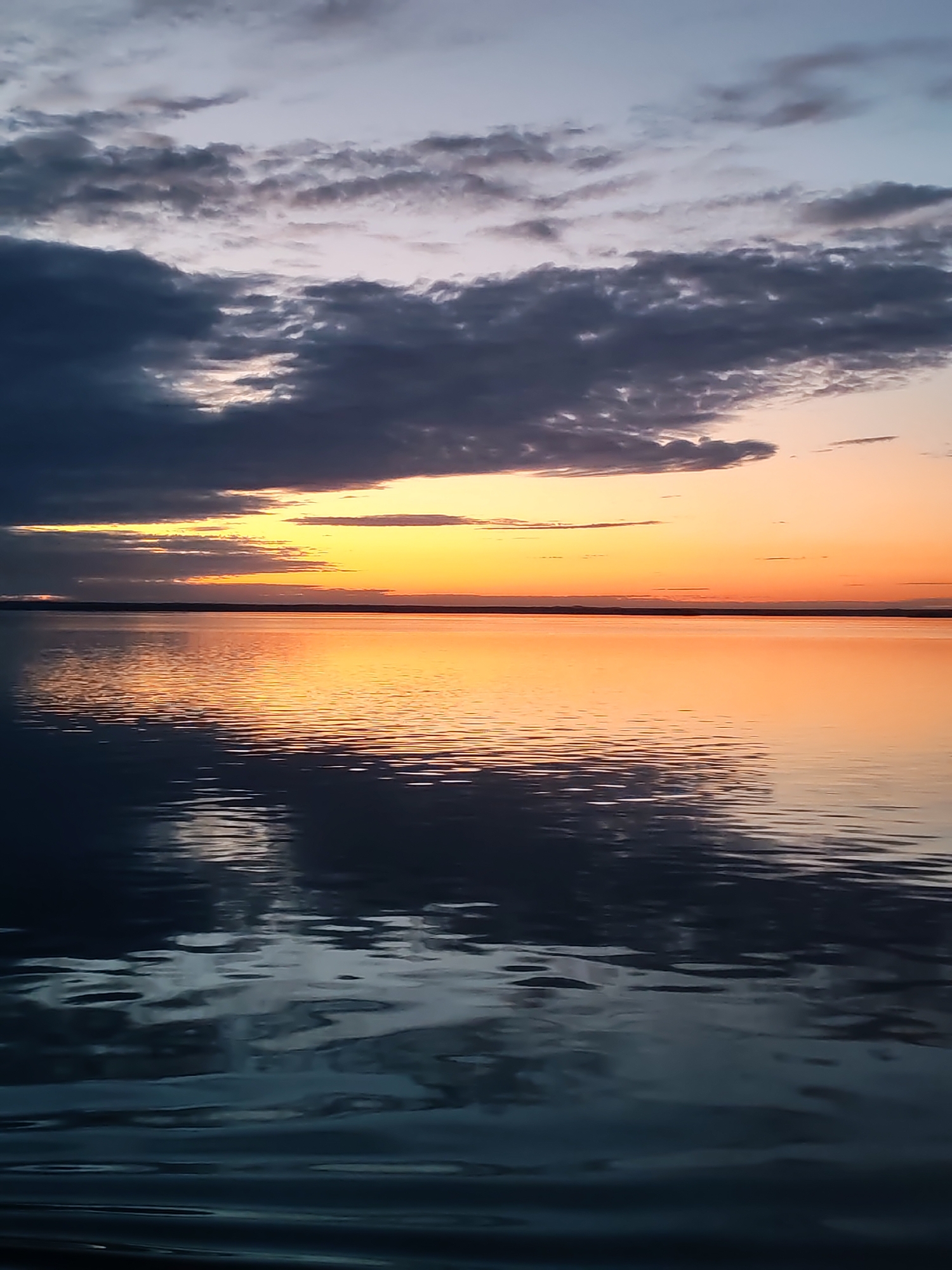 The image depicts a serene sunset or sunrise over a vast body of water, likely a lake or a wide bay. The sky is dominated by dramatic clouds, transitioning from brilliant yellows and oranges at the horizon, where the sun is located, to pale blues and greys higher up. Dark, heavy clouds are prominent in the upper left, casting deep shadows and partially obscuring the light source, while lighter, wispy clouds are scattered elsewhere.
The water's surface acts as a mirror, reflecting the vibrant colors of the sky. A broad path of intense orange and yellow light shimmers across the central portion of the water, indicating the sun's reflection. Away from this direct reflection, the water appears a deeper blue-grey, but is still textured with numerous ripples and gentle waves that create intricate patterns of light and shadow across its surface. A faint, dark line stretches horizontally across the distant horizon, suggesting a low-lying landmass or a distant shore.
No human subjects, specific man-made objects, or discernible text are visible, emphasizing the natural landscape. The scene conveys a sense of tranquility and the grandeur of nature at dawn or dusk.