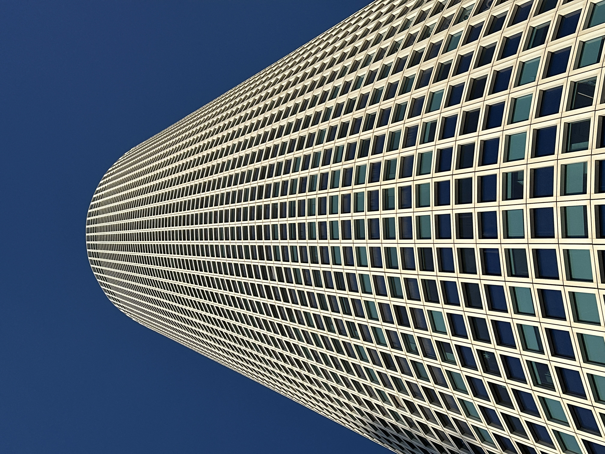 This is an abstract architectural photograph captured from a low angle, looking upwards at a large, modern building against a clear, deep blue sky. The building dominates the frame, its curved facade extending diagonally from the bottom right towards the top left corner. The primary subject is the building's exterior, which is composed of a dense grid of uniform square or rectangular window panels, each set within a light-colored, possibly white or off-white, frame. The windows themselves appear to be dark-tinted or reflective, showing varying shades of blue and sometimes hints of interior darkness or reflections.
The distinct curvature of the building suggests a cylindrical or elliptical shape, creating a sweeping diagonal line across the composition. The repetitive pattern of the windows, combined with the building's curve, creates a sense of depth and scale. The bright and even lighting indicates daytime, likely mid-day, under clear, sunny weather conditions, with no clouds visible in the stark blue sky. There are no people or discernible activities taking place. The overall impression is one of modern design, precision, and urban grandeur. No text is visible in the image. The building is located in Tel-Aviv, Israel.