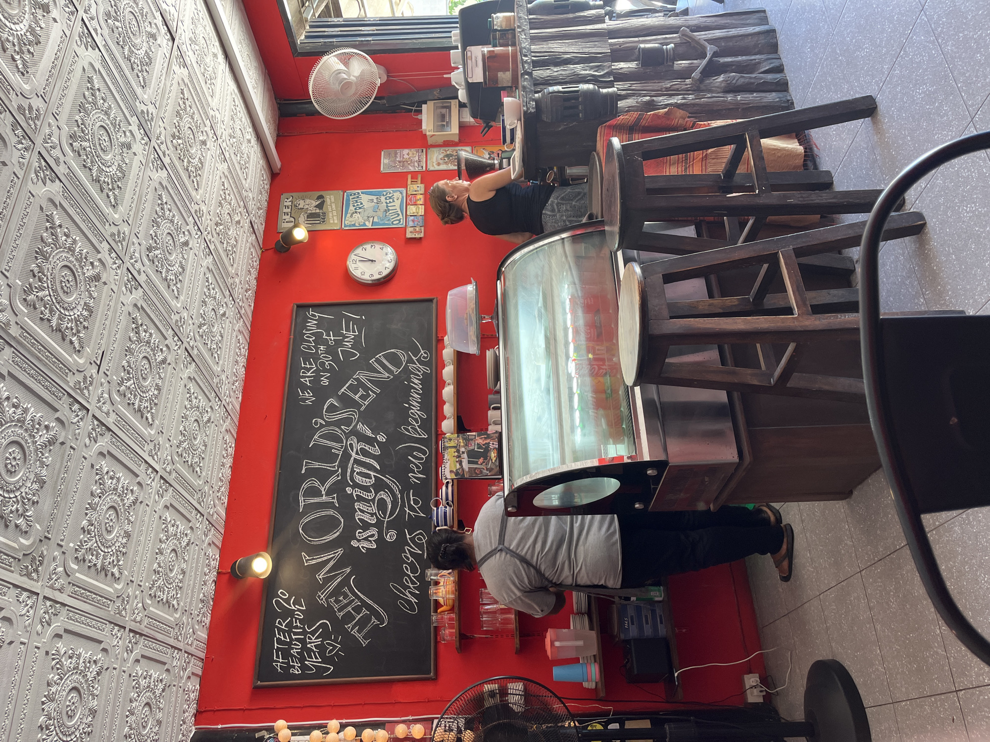 The image depicts the interior of a cafe in ตำบลเกาะพะงัน, Thailand. The cafe has bright red walls and a highly ornate tin ceiling. A large chalkboard mounted on the wall features handwritten messages, including "AFTER 20 BEAUTIFUL YEARS THE WORLD IS NIGH! WE ARE CLOSING ON 30TH OF JUNE! Cheers to new beginnings." Several framed posters are also displayed on the wall.
In the foreground, a refrigerated display case is visible, filled with drinks. Two wooden bar stools are positioned in front of it. A woman with dark hair and an apron is standing behind the counter, facing away from the camera, appearing to work. To the right, another woman with blonde hair, wearing a black tank top and patterned skirt, is serving at the counter. The counter itself is rustic in appearance, with wooden elements and a checkered fabric draped over it.
Various shelves on the red wall hold stacks of plates, cups, and a teapot. A modern clock is mounted on the wall, along with a small white fan. In the lower left corner, a large black floor fan is present. The floor is tiled. The overall impression is of a casual, perhaps slightly bohemian, establishment.